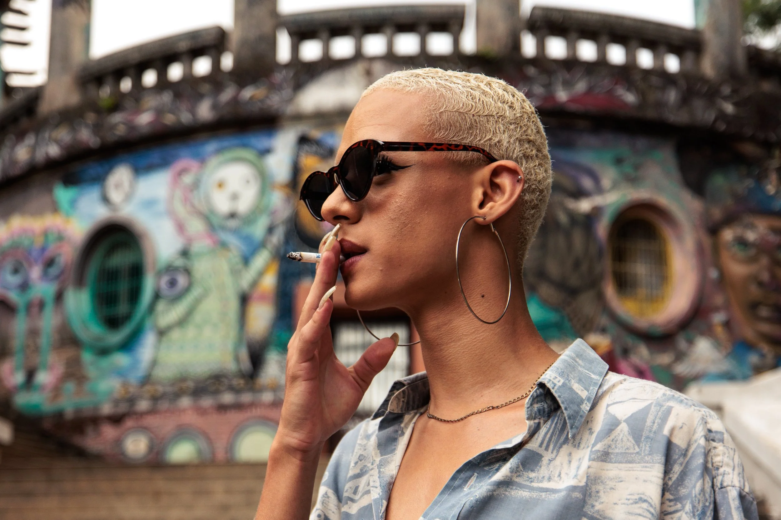 A person with short platinum blonde hair wearing dark sunglasses and hoop earrings holds a cigarette near their lips. They are dressed in a patterned button-up shirt and have two earrings in one ear. The background features a colorful graffiti mural 