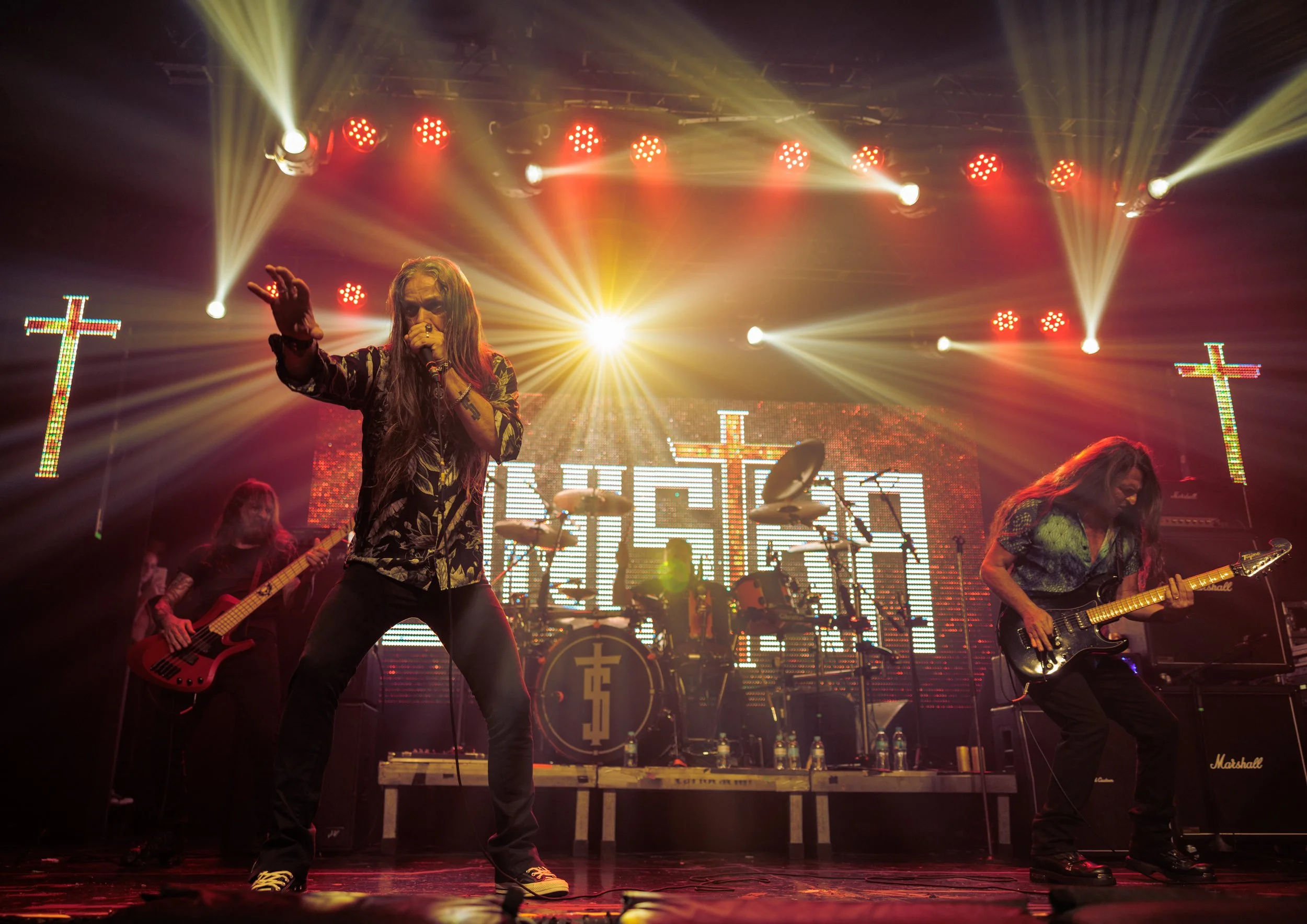Musicians perform on stage with colorful lighting, including a singer in the center holding a microphone, guitarists on either side, and a drummer in the background. Bright lights and illuminated crosses are visible in the background.