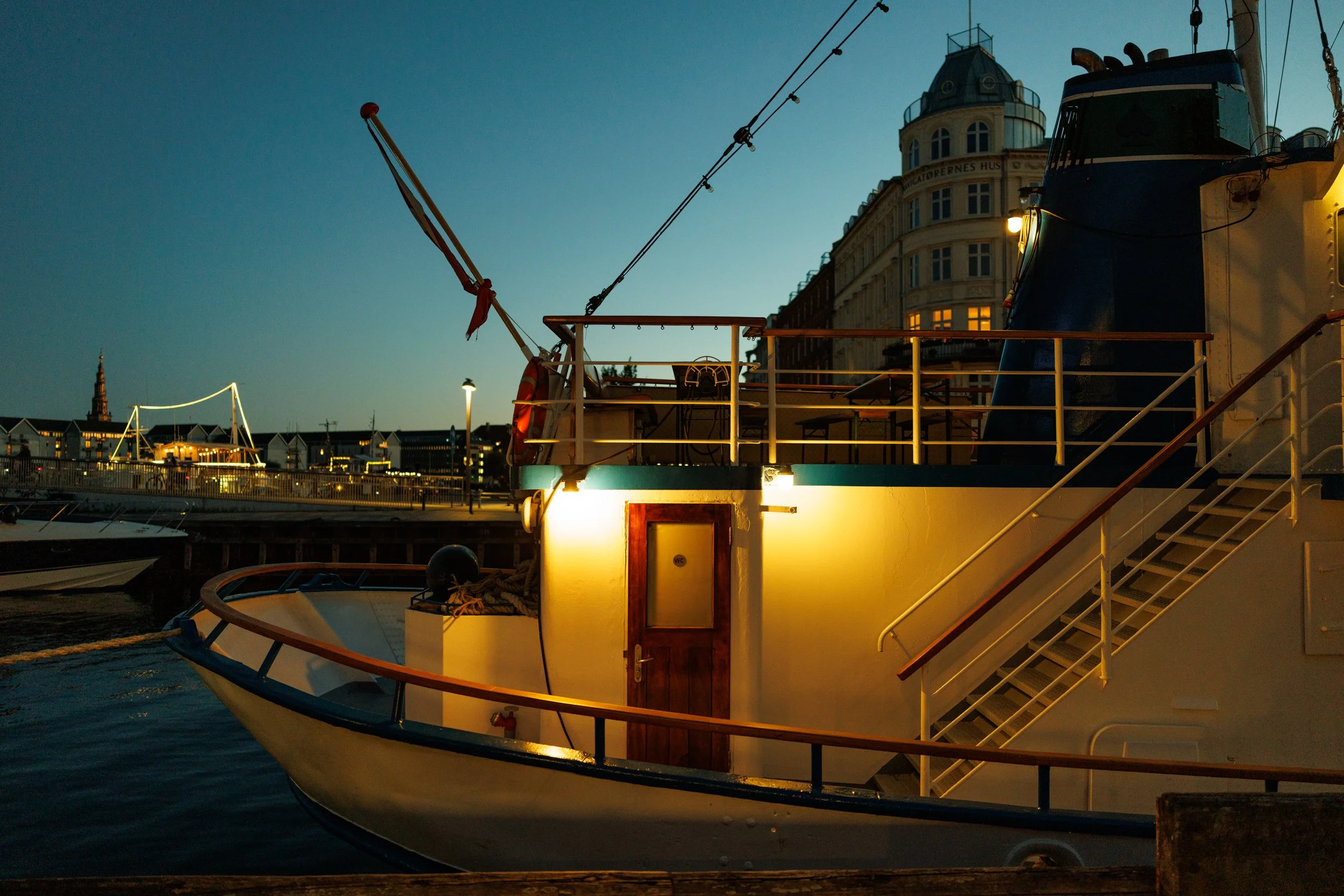 A docked boat at sunset with a cityscape in the background, including historic buildings and a church spire, illuminated by streetlights.
