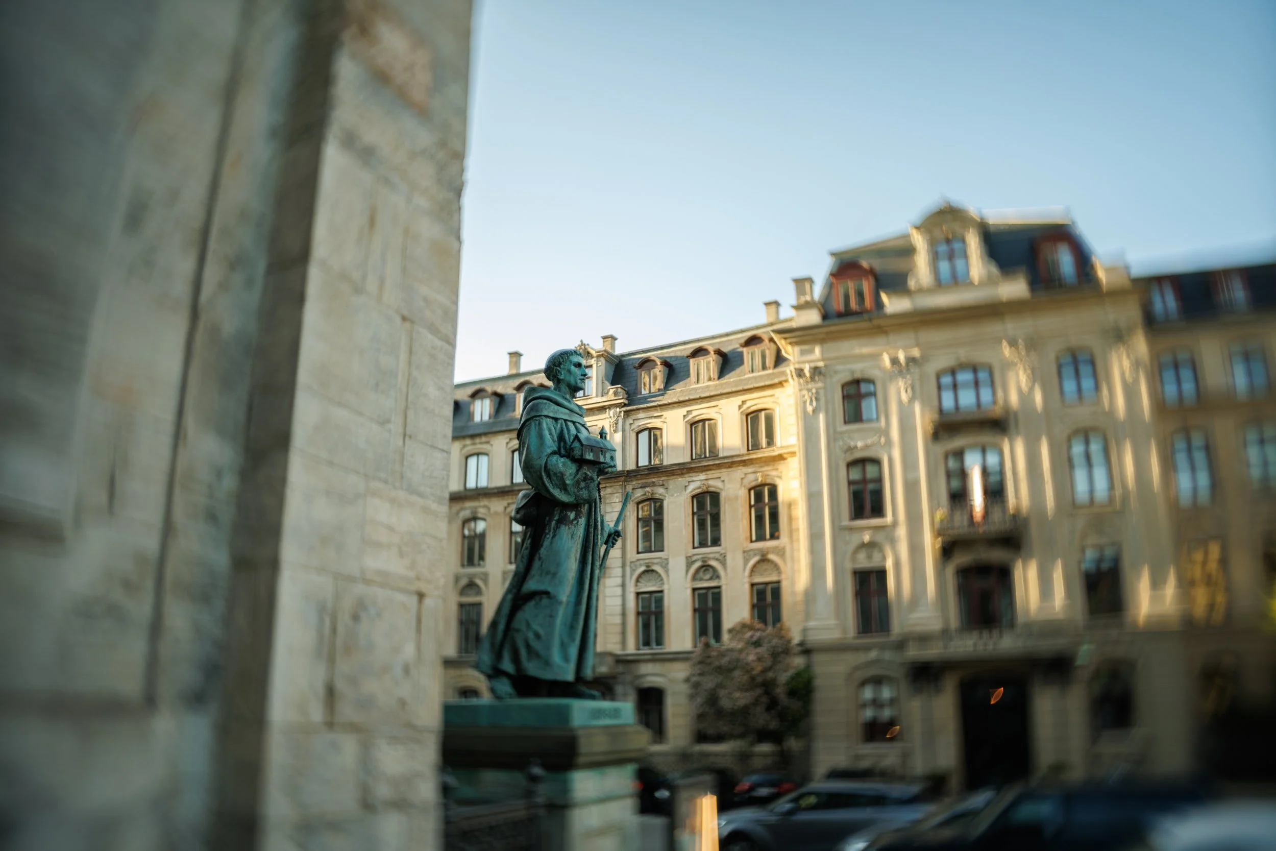 A bronze statue of a man in historical attire holding a book and a scroll, situated in a city square with classical European-style buildings in the background under a clear blue sky.