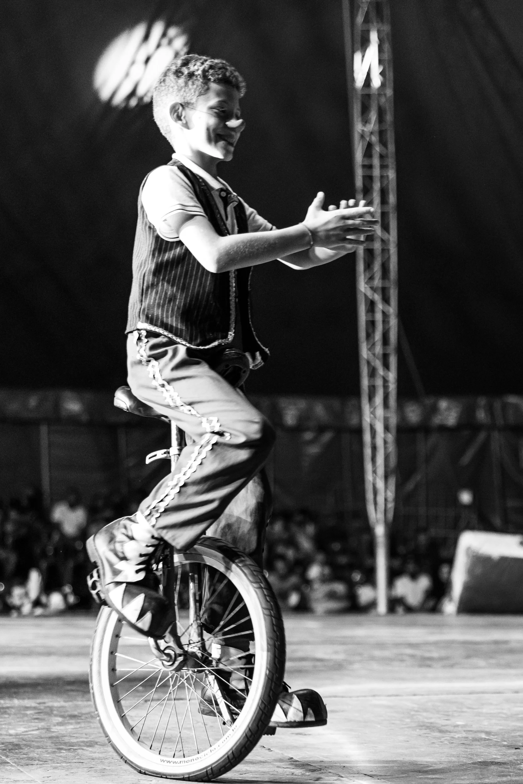 A young person performing a BMX stunt on a bike in an indoor arena, with audience in the background, black and white photo.