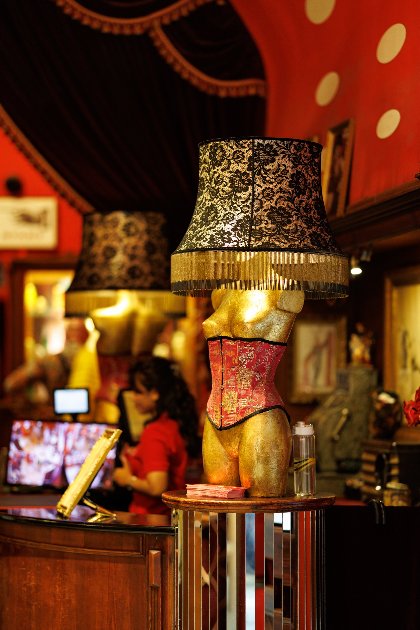 Decorative lamp shaped like a female torso with a lace lampshade, in a dimly lit room with red walls and people in the background.