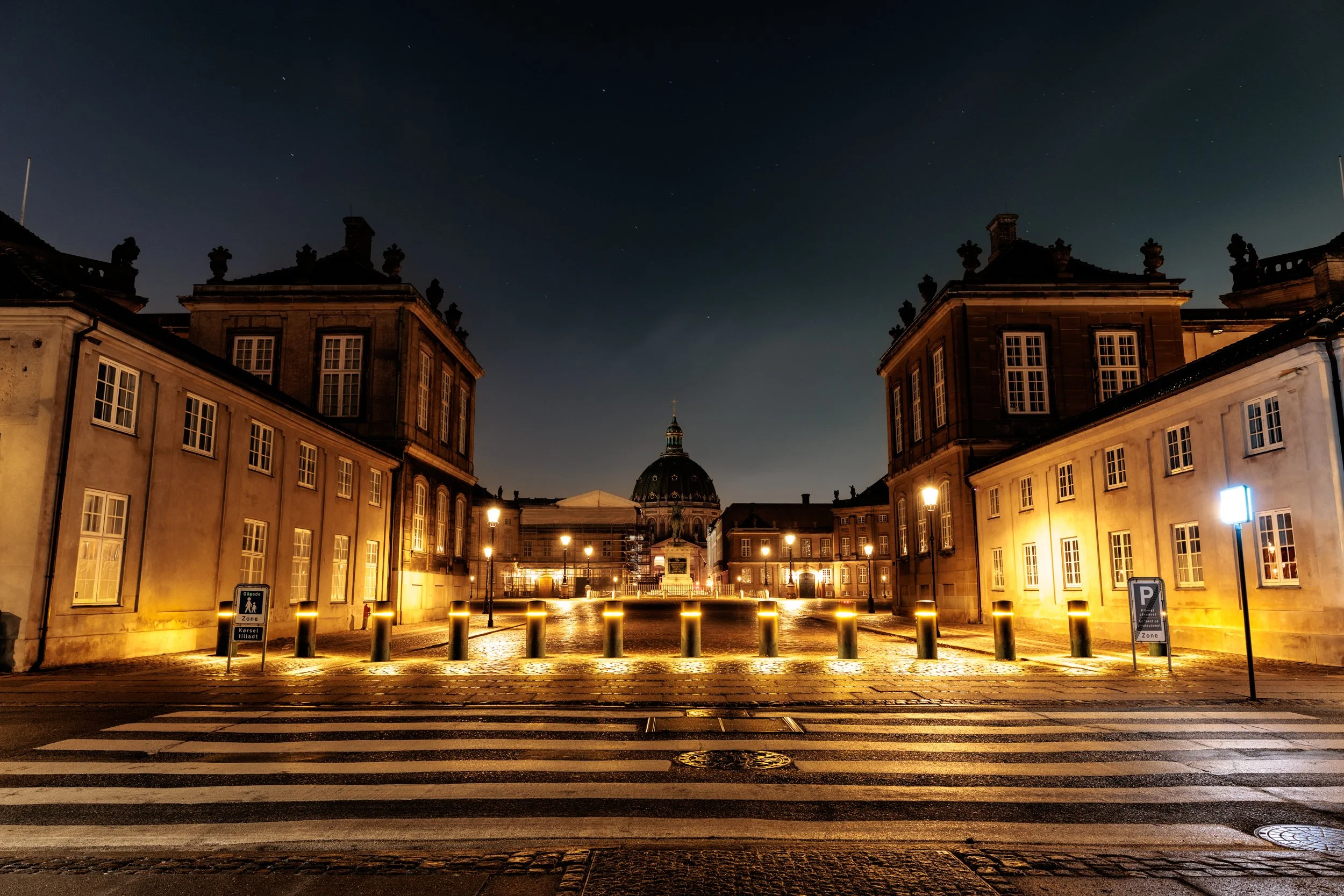Nighttime view of historic buildings with illuminated windows and street lamps, a cross and dome in the background, cobblestone street with pedestrian crossing, and signs indicating parking and zone restrictions.