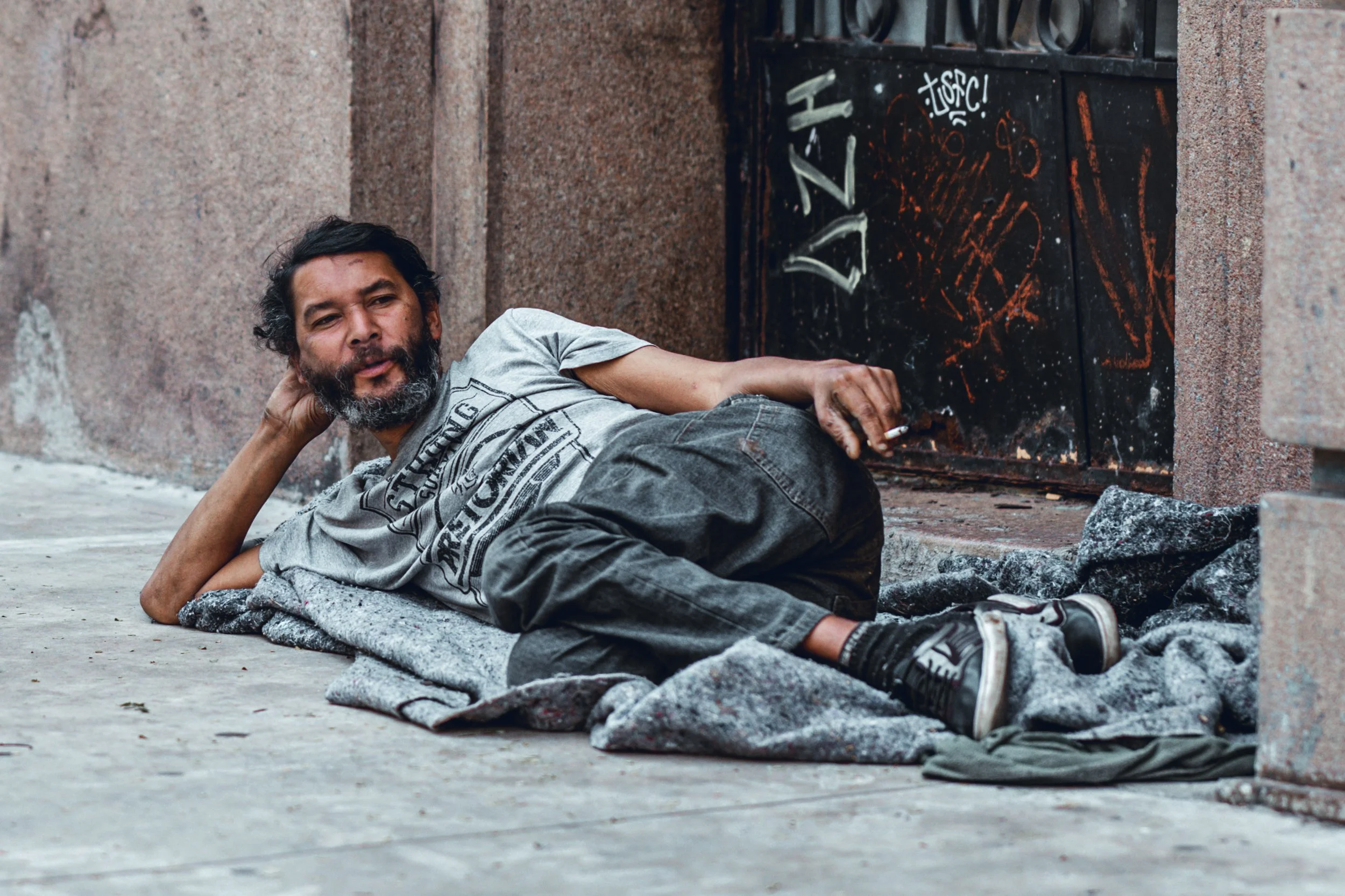 A man with dark hair and a beard lies on the sidewalk, resting his head on his hand near a graffitied metal box, against a brick wall.