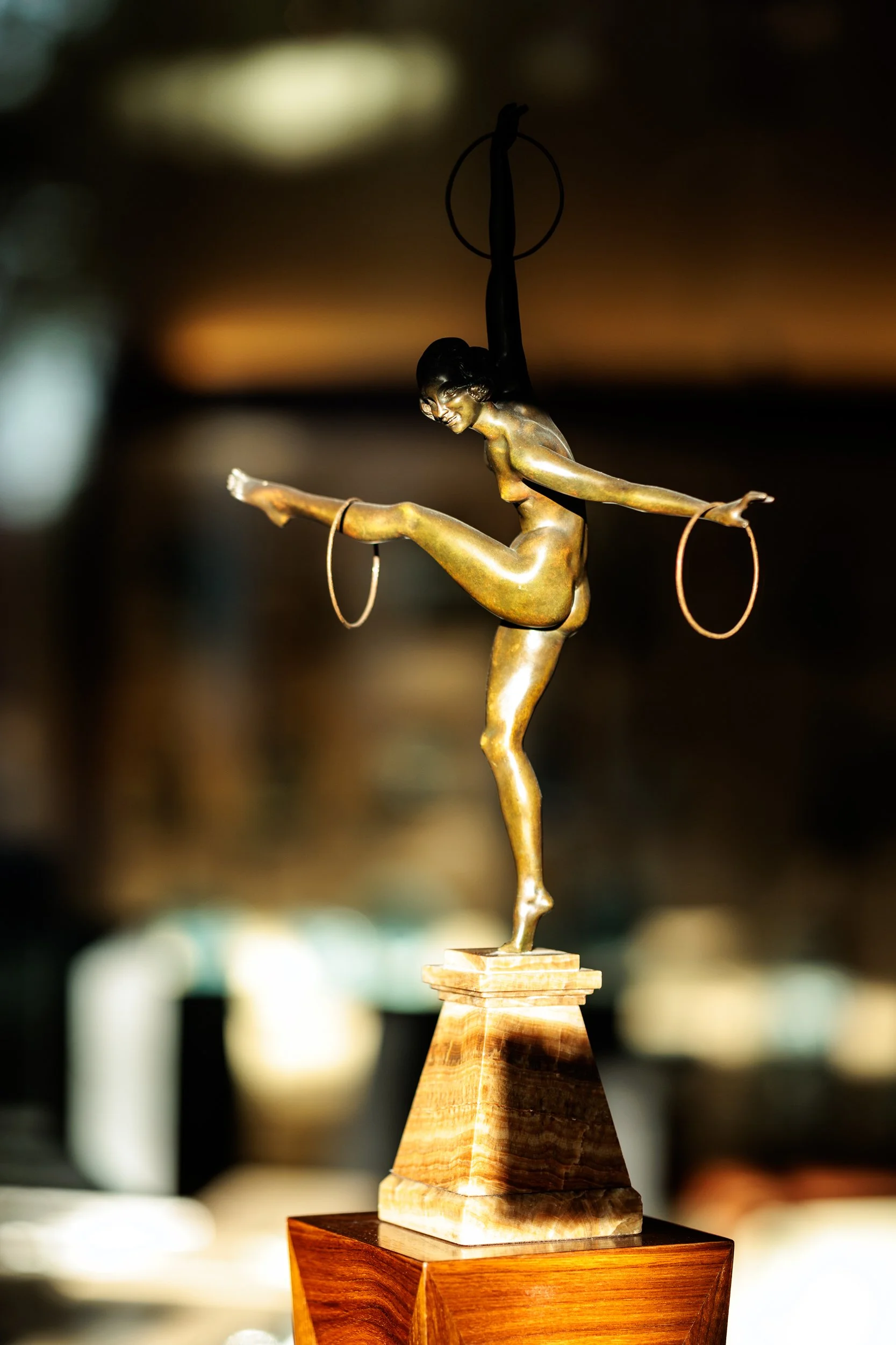 A gold-colored sculpture of a dancer on a wooden base, with one arm extended upward holding a ring, and the other arm extended outward holding a ring, in a ballet pose.