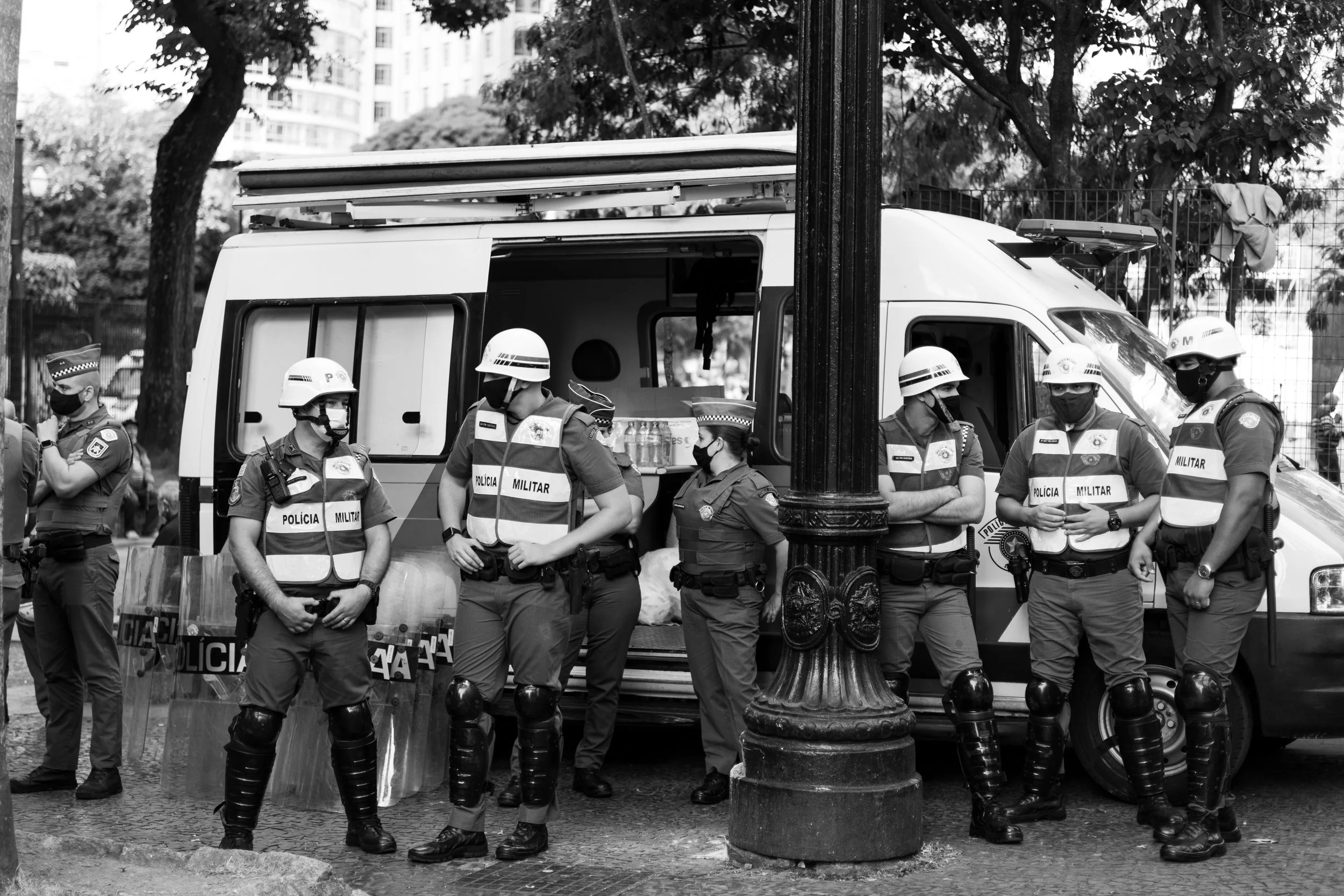 Black and white photo of several police officers in uniform and helmets standing in a line behind a street barricade in an urban park area, with trees and buildings in the background.