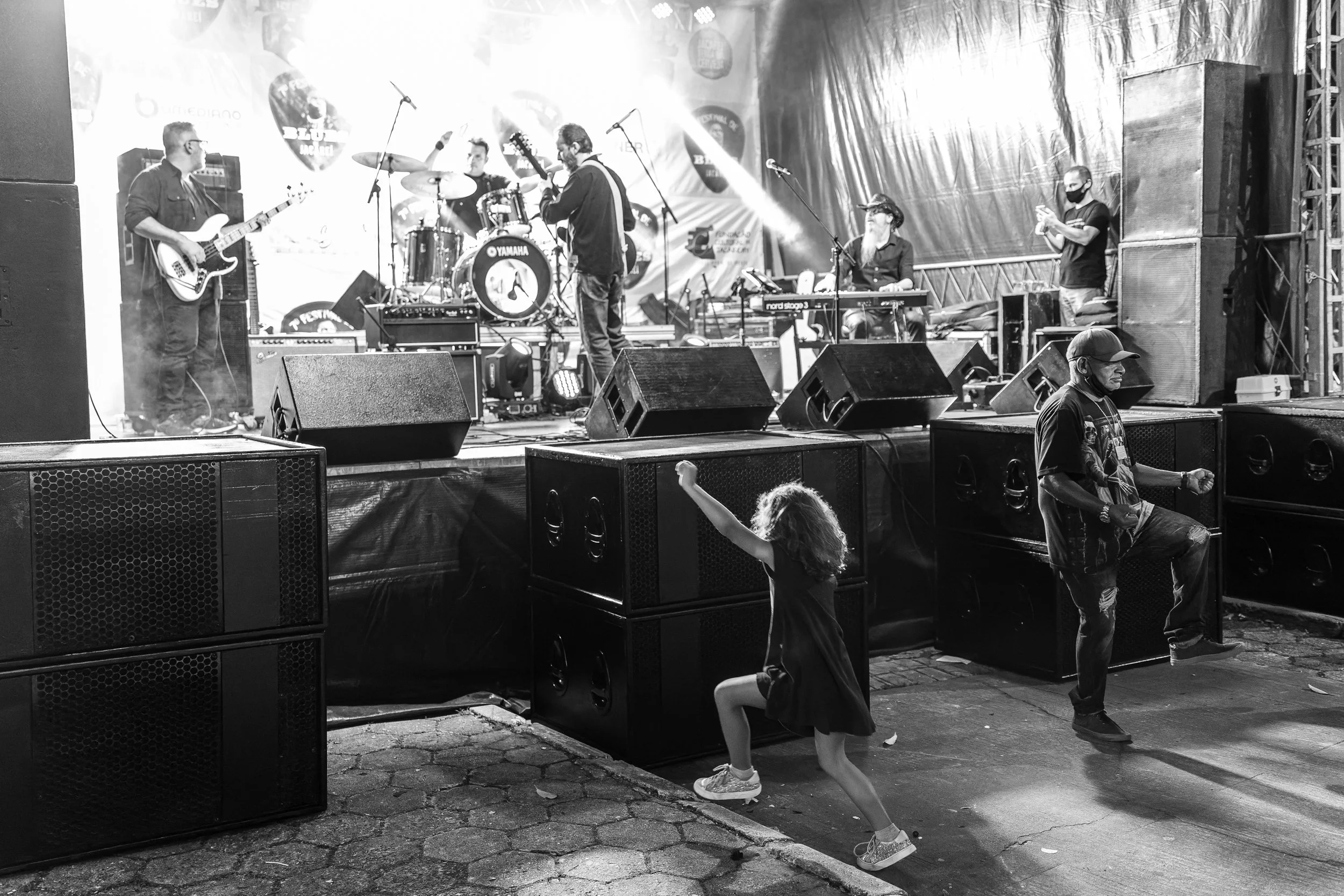 A black and white photo of a live band performing on an outdoor stage at night. The band includes a guitarist, drummer, keyboardist, and a vocalist. Two people in the audience are dancing near the stage, with one person wearing a mask and a hat, and 