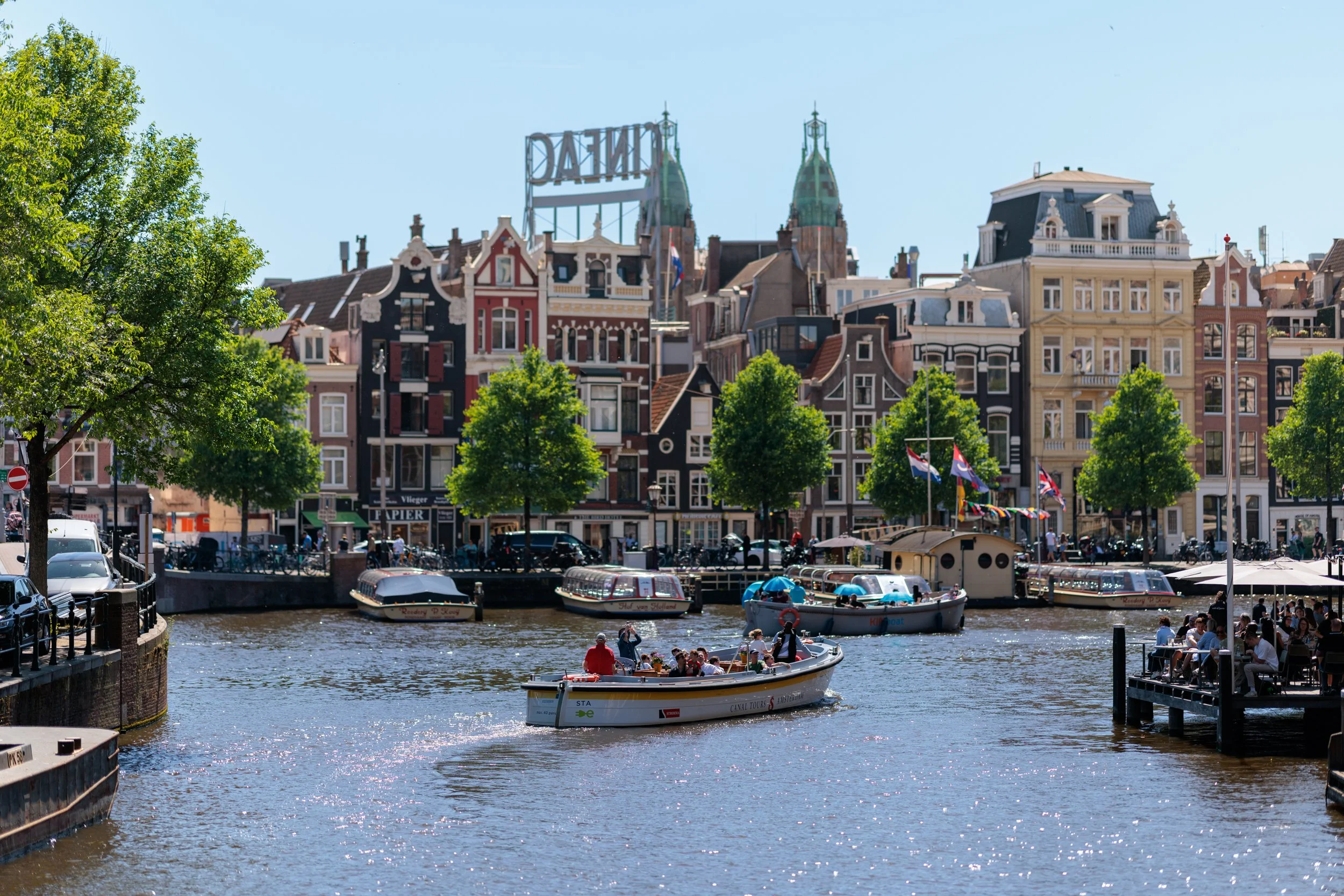 Canal with boats and people walking along the waterfront, featuring historic European-style buildings with ornate facades and rooftops, and lush green trees lining the area, under a clear blue sky.