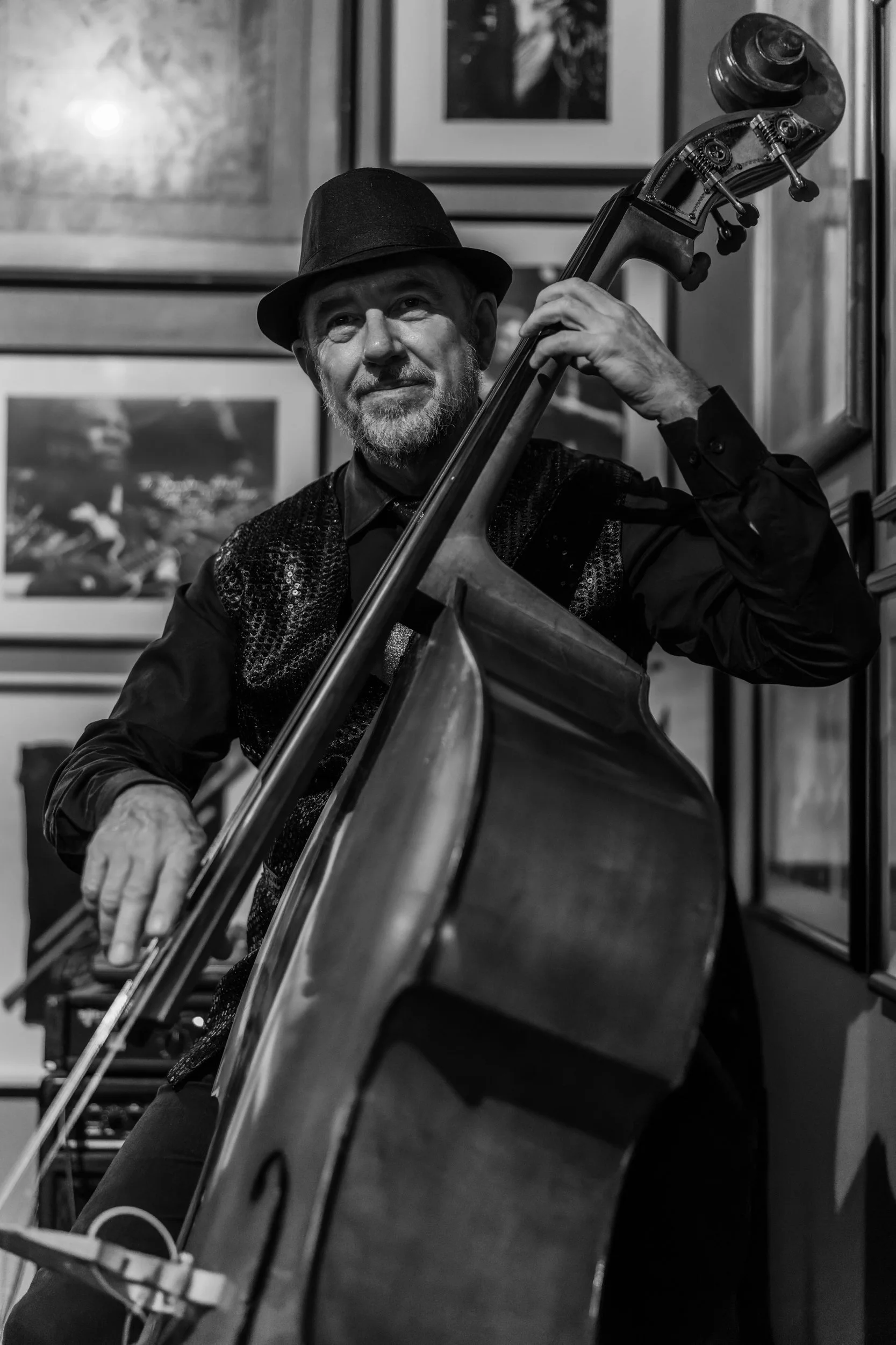 A man with a beard and mustache playing a double bass, wearing a fedora hat and dark clothing, in an art gallery with framed pictures on the wall.
