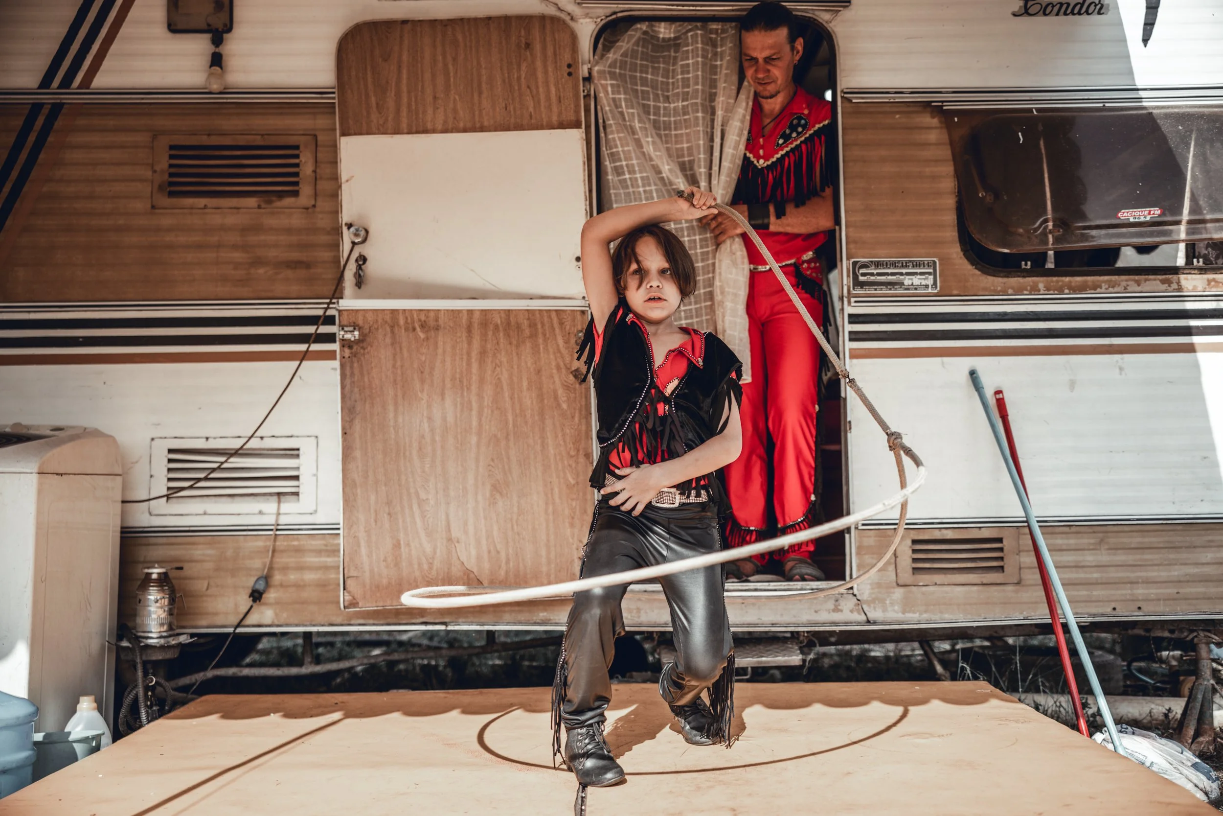 A woman with dark hair and leather pants dancing on a stage outside a wooden camper trailer, with a man in a red shirt watching from the door.