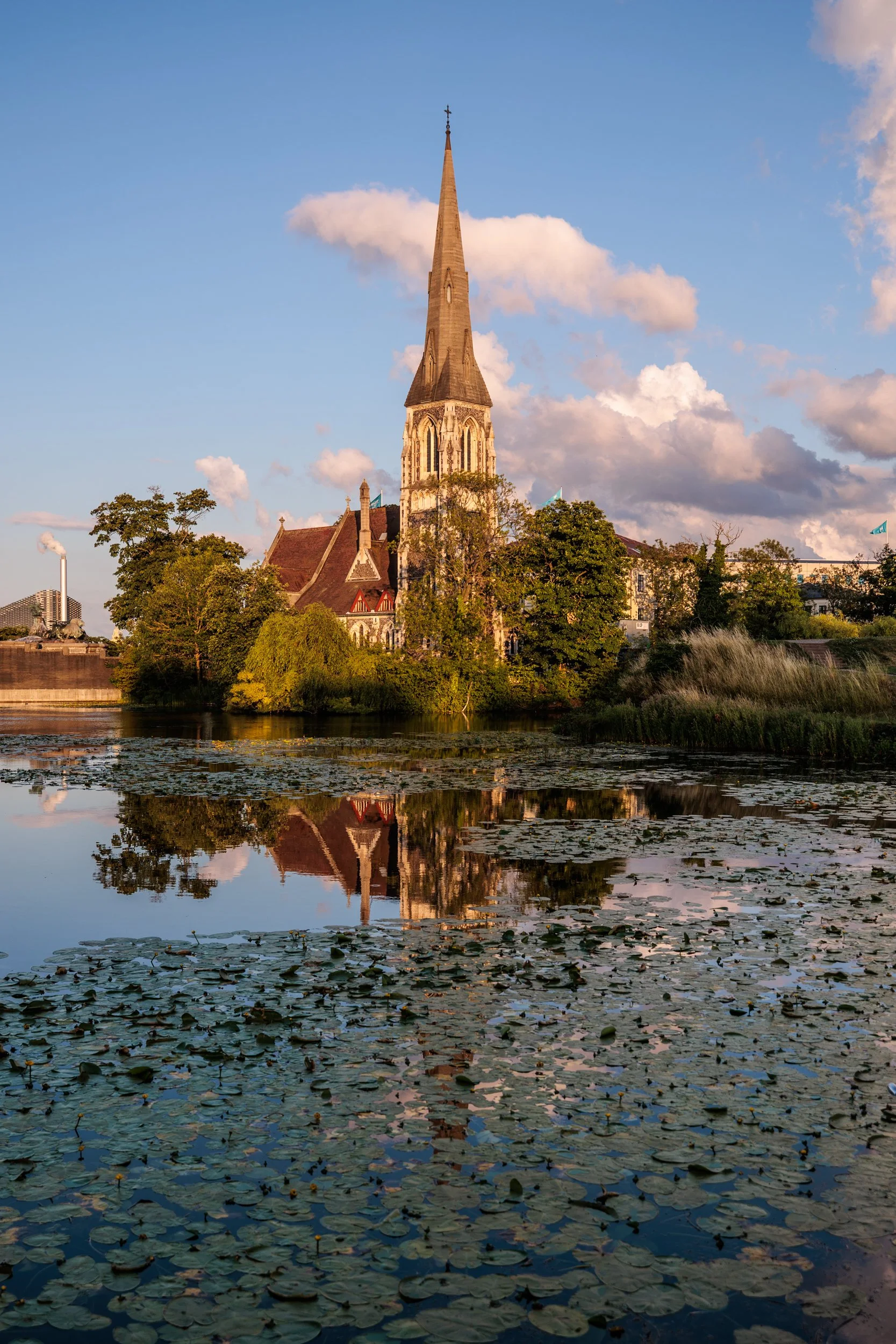 A church with a tall steeple and Gothic architecture, surrounded by trees and overlooking a body of water with lily pads. The sky is partly cloudy with the sun setting, casting a warm glow.