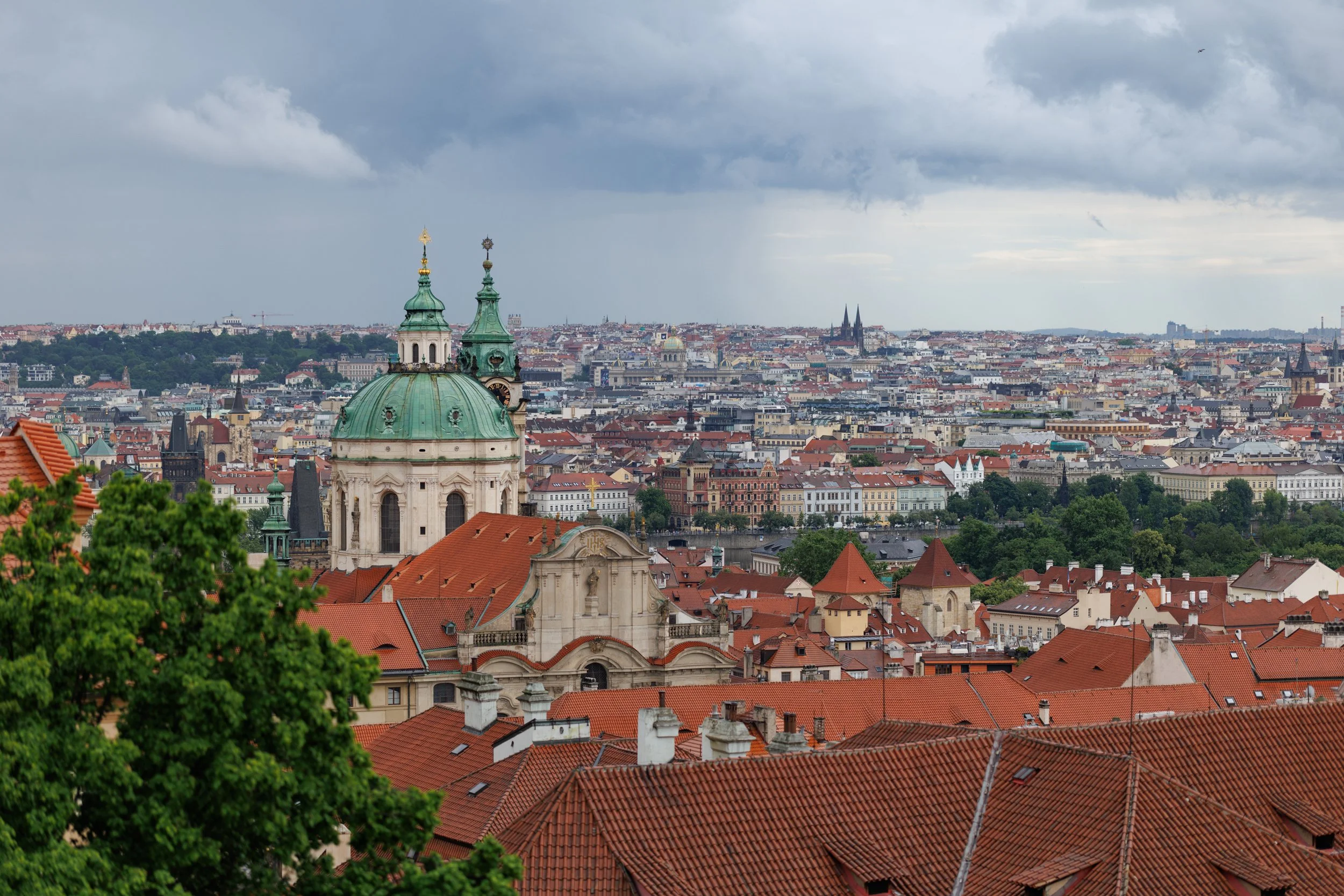 View of Prague cityscape with historic buildings, red rooftops, church spires, and cloudy sky.