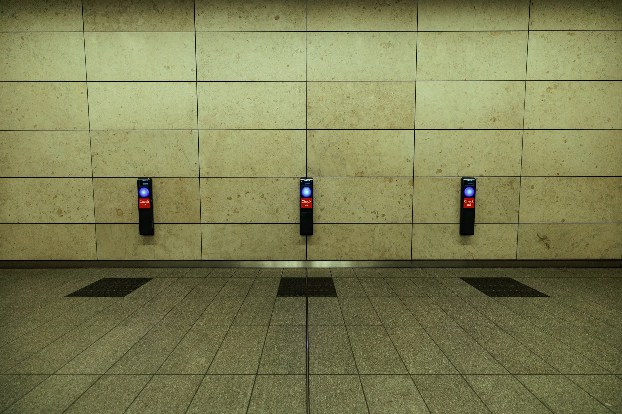 Three floor-standing red and black security check-in devices with blue glowing buttons, mounted on a beige tiled wall in an airport or train station.
