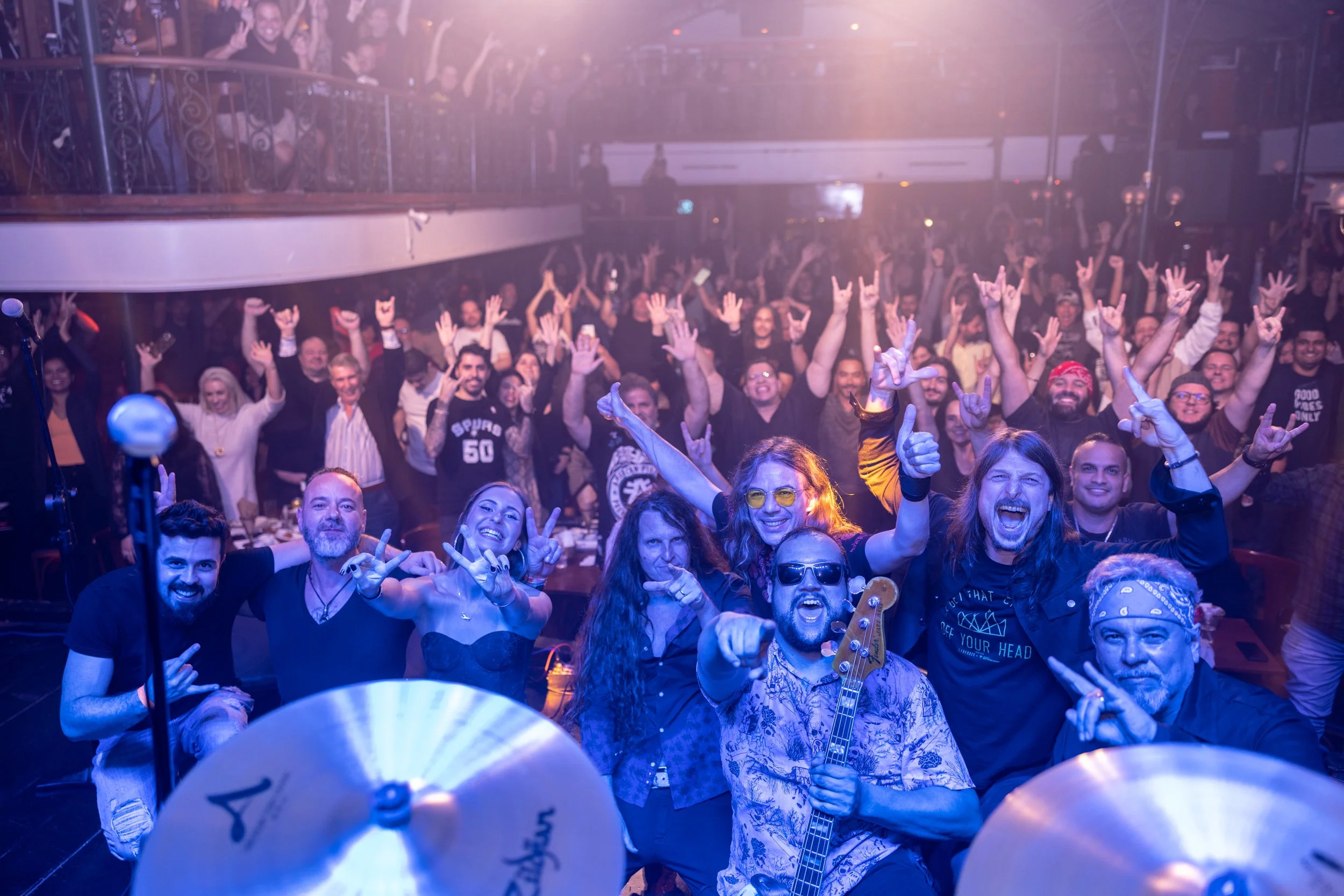 A large crowd of people at a concert or party, smiling, raising their hands, and making hand gestures. The atmosphere is lively with purple and pink lighting, and some people are holding musical instruments, indicating a celebration or musical event.