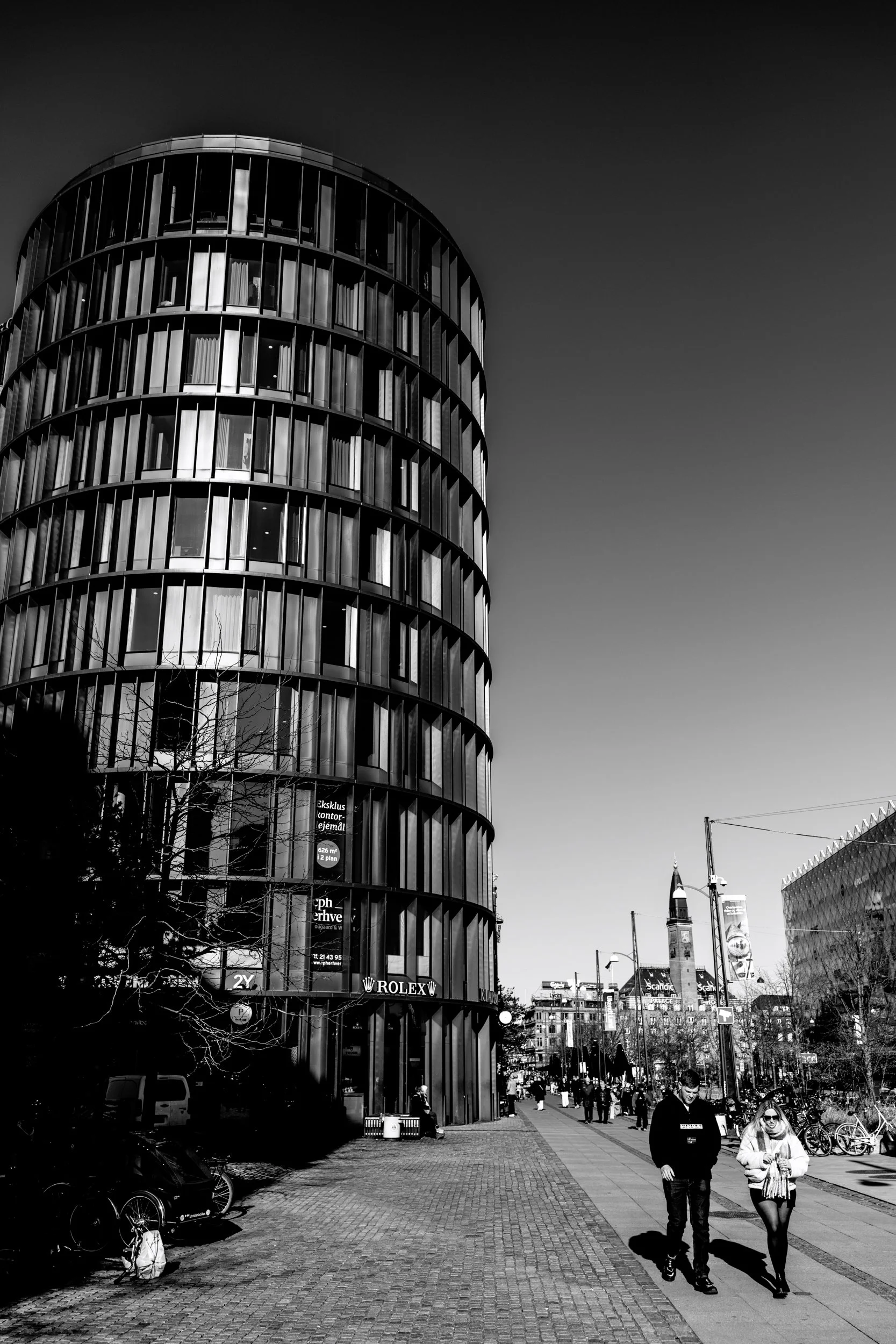 Black and white photo of modern cylindrical building with glass windows on an urban street. Pedestrians walk along the sidewalk, some carrying bags, with bicycles parked nearby. In the background, a historic clock tower is visible.