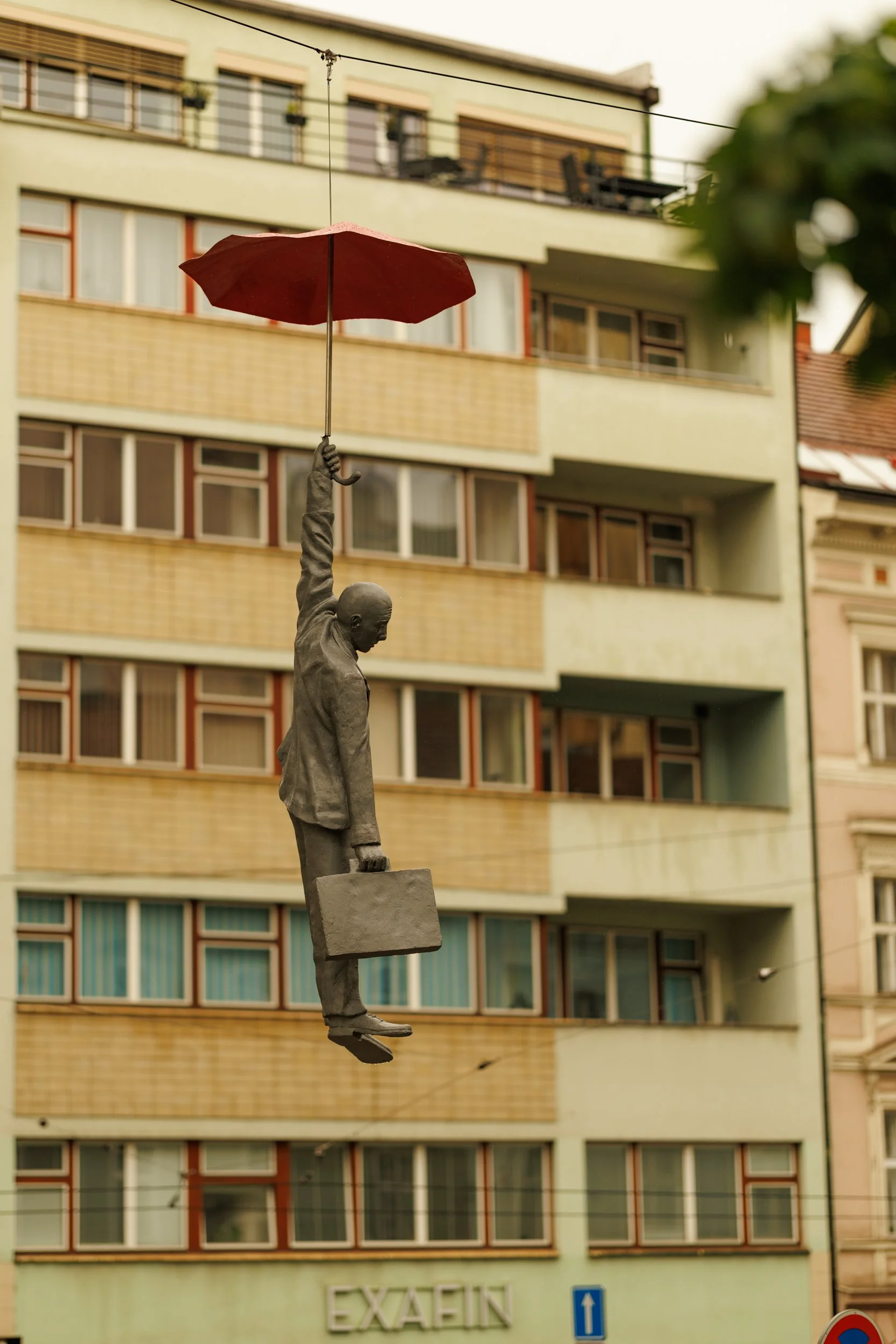A statue of a man holding a red umbrella above his head, suspended in mid-air, in front of a multistory building with several windows and balconies.