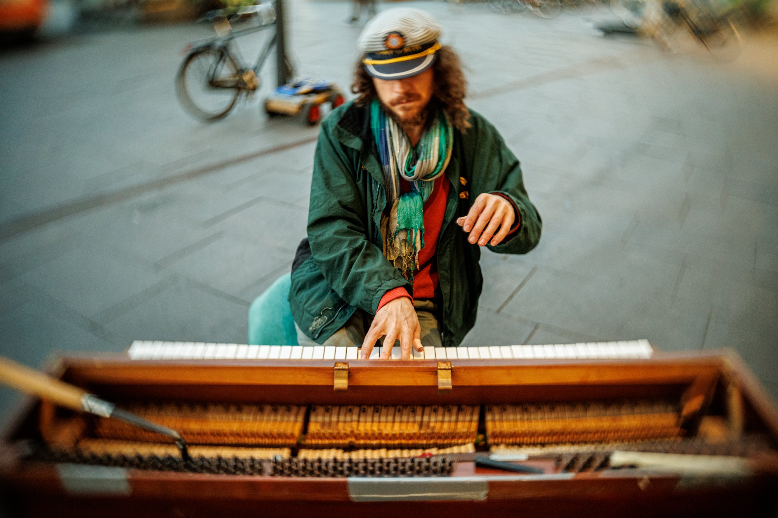 A man with curly hair and a beard, wearing a captain's hat and green jacket, is playing a piano outdoors. A bicycle and stroller are in the background, along with blurred pedestrians.