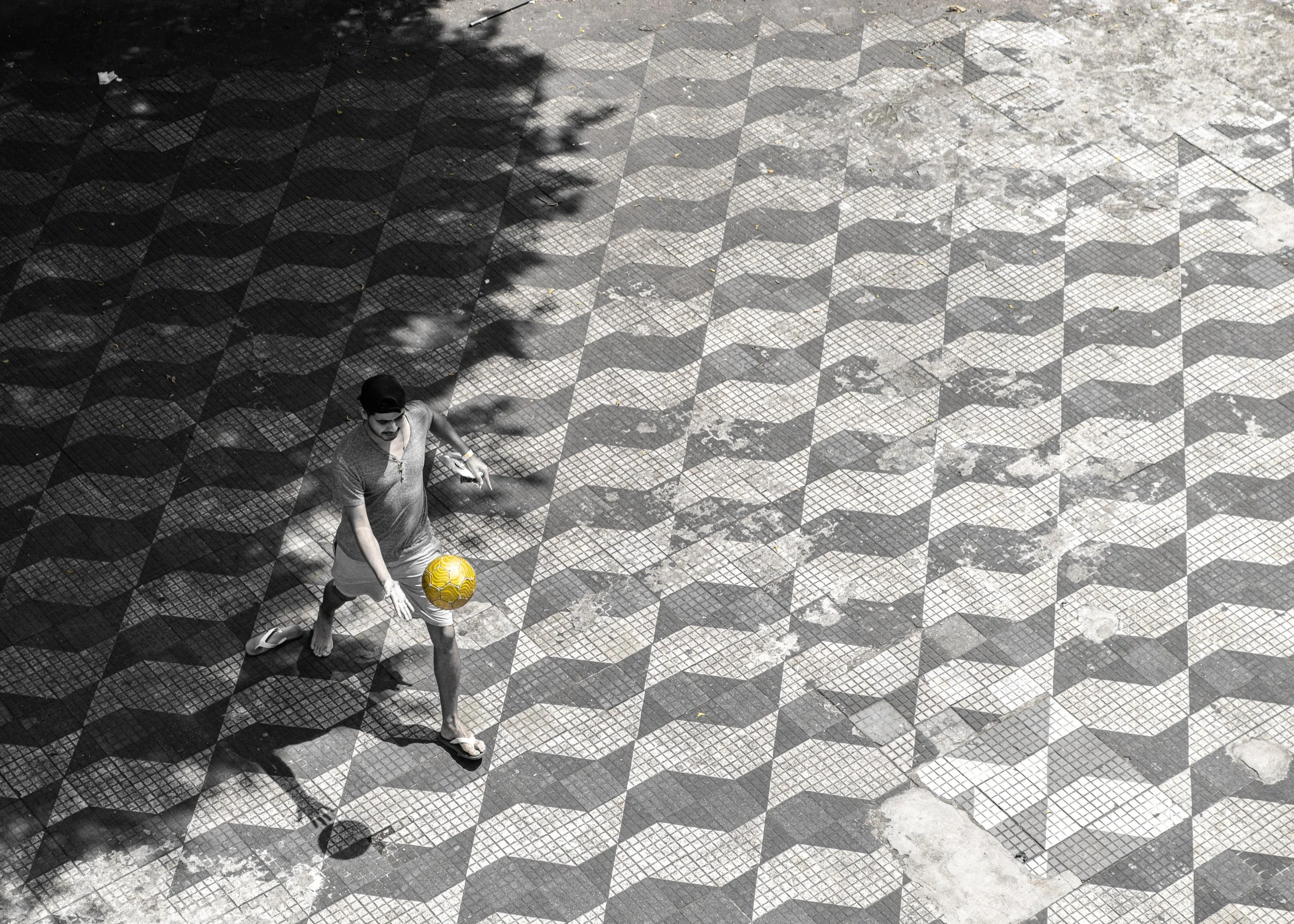 A young man wearing a cap and shorts playing soccer outdoors on a patterned tile surface, with shadows of nearby trees cast on the ground.