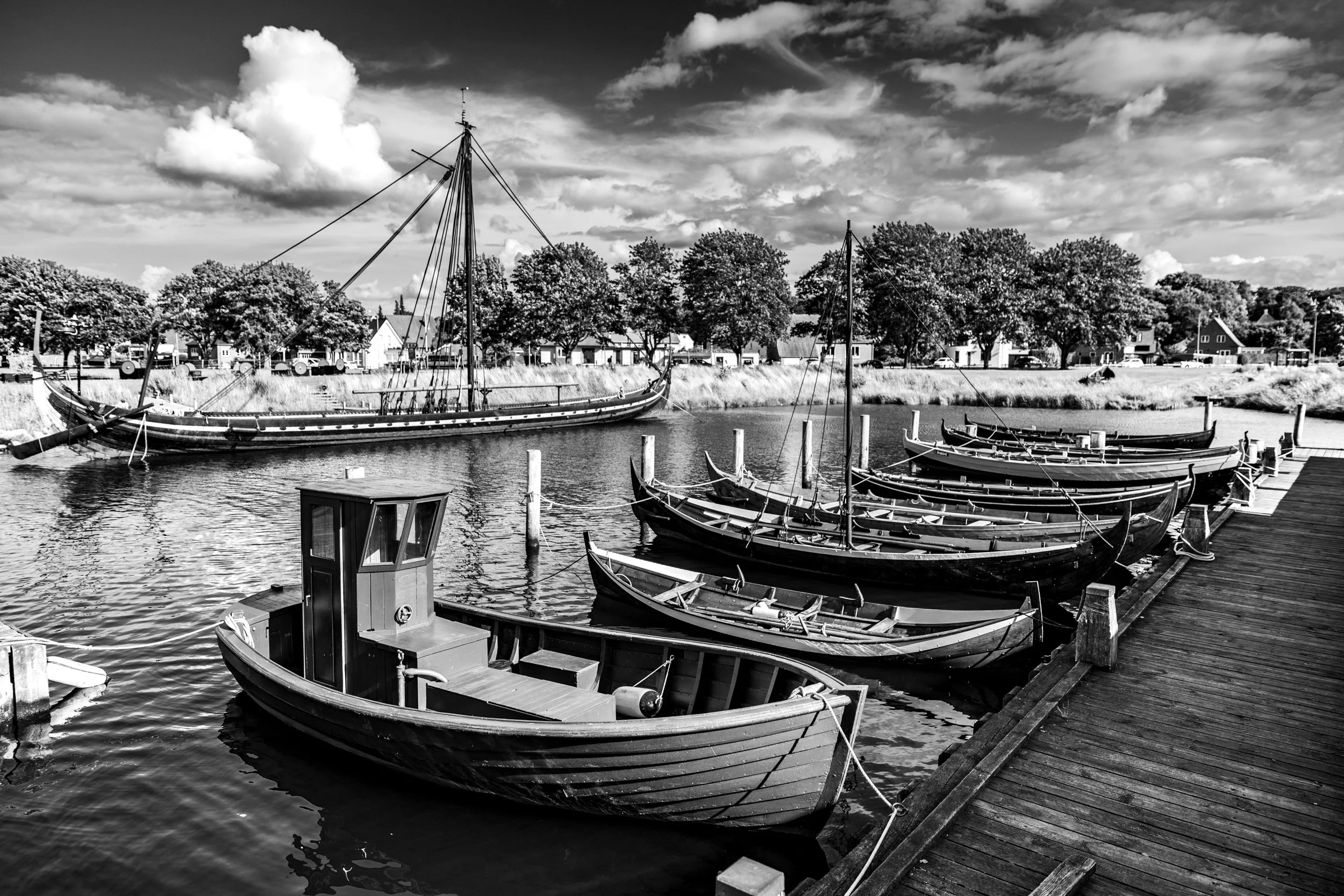 Black and white image of boats docked along a river with houses and trees in the background and a partly cloudy sky overhead.