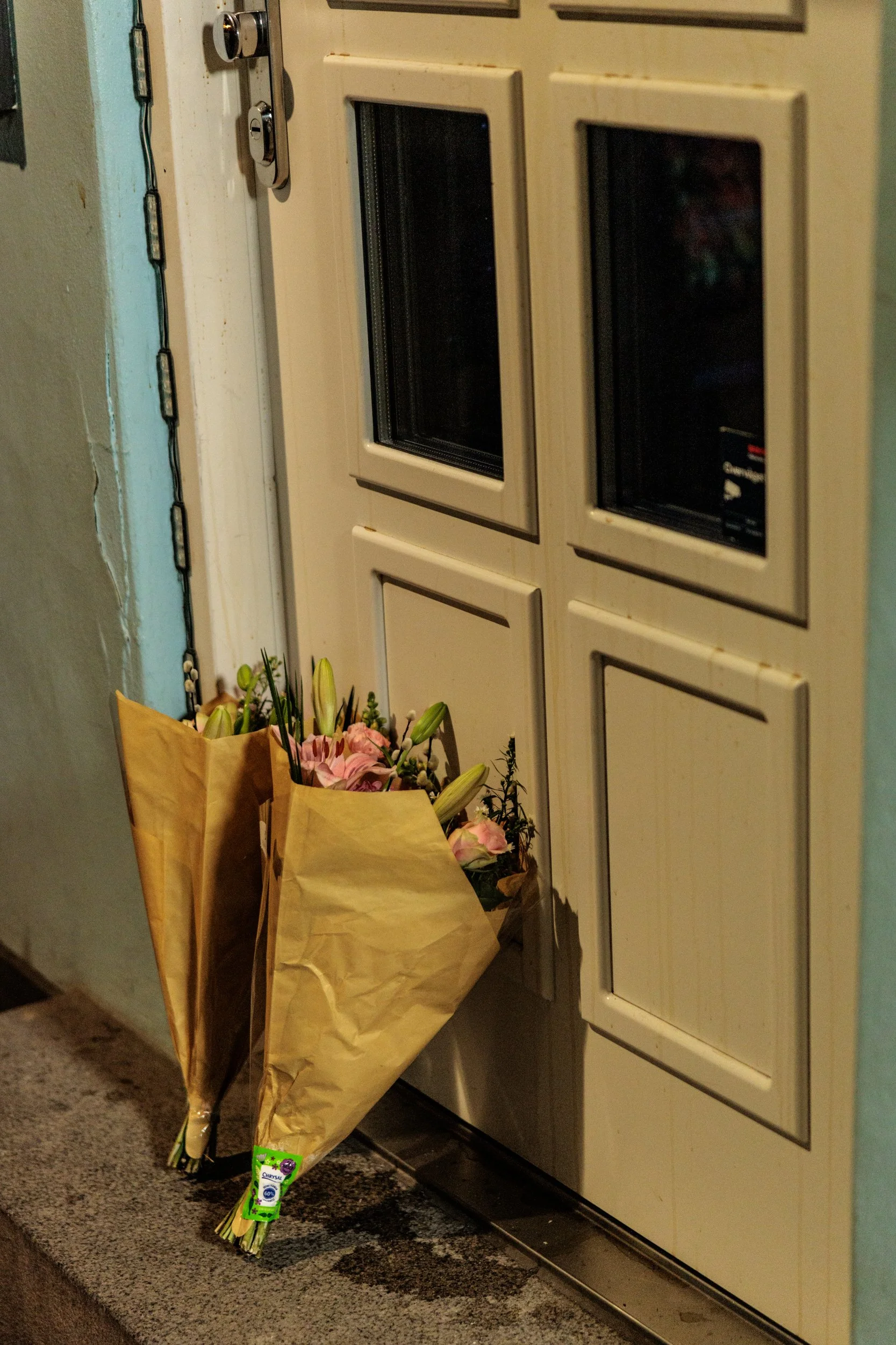 Bouquet of flowers wrapped in brown paper and a tube of hand cream placed at the bottom, positioned near a door with a mail slot. The scene appears inside a building entrance or hallway.