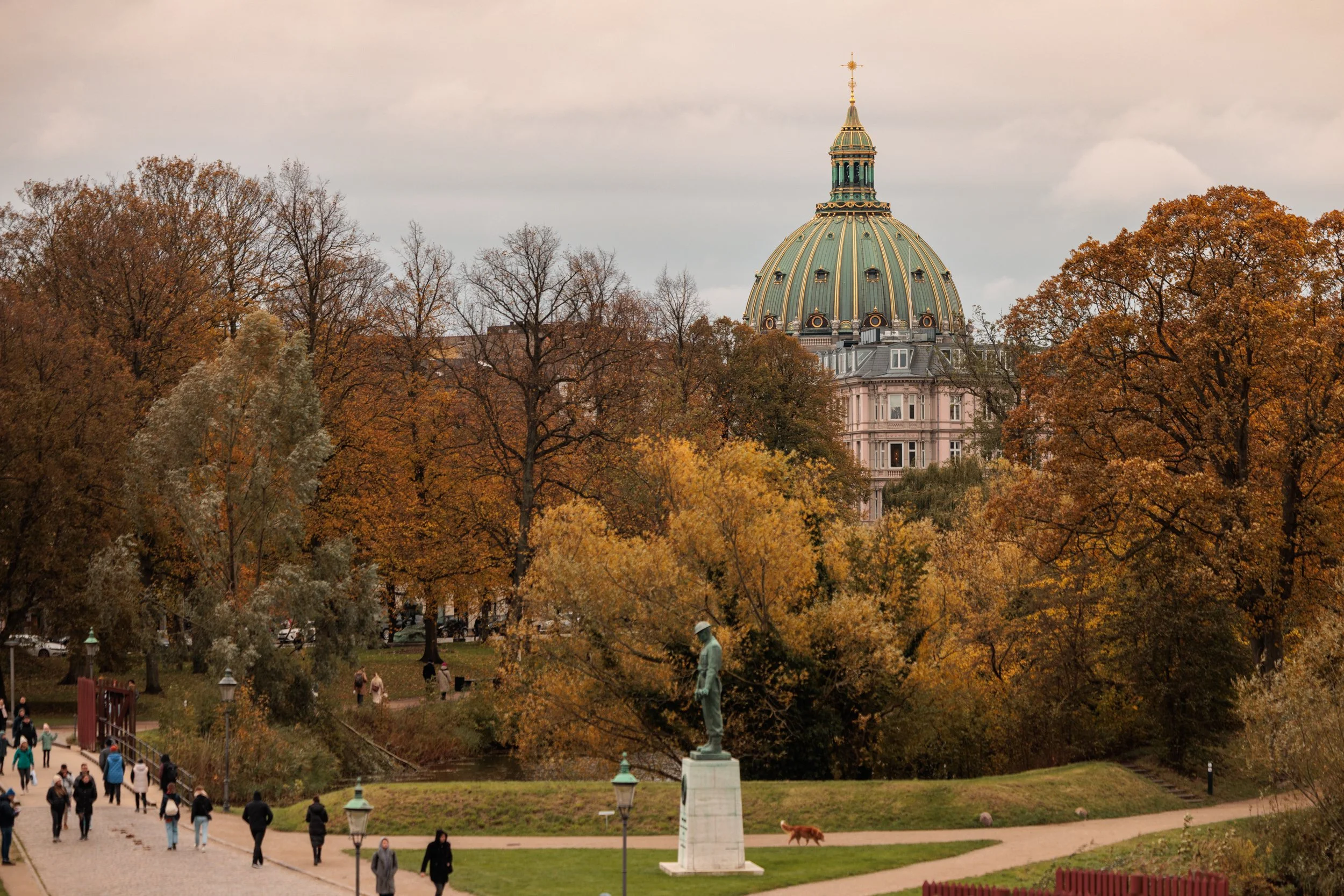 View of a park with people walking, trees with autumn leaves, a white statue on a pedestal, and a building with a gold-domed roof in the background.
