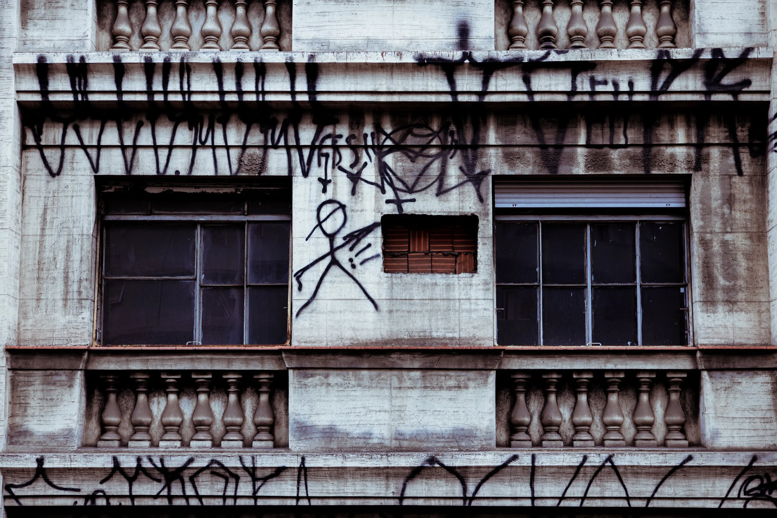 A weathered building facade with graffiti spray paint, including a pentagram and various tags, two broken windows with darkened panes, and a partially boarded-up window.