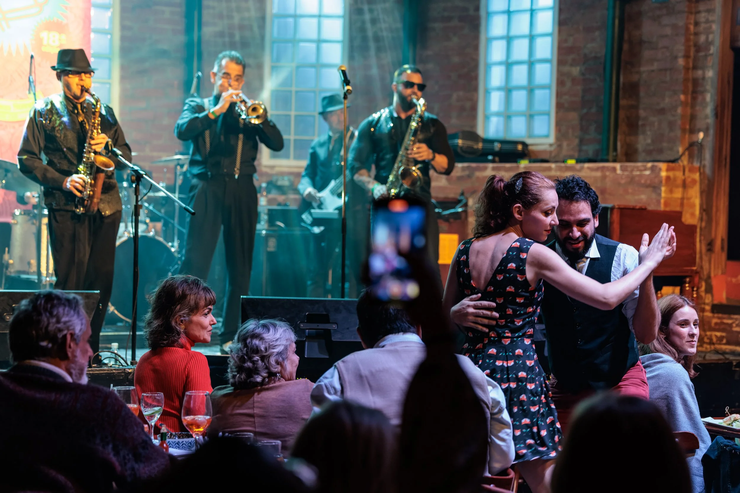 Live jazz band performing on stage at a lively indoor event with audience mingling and dancing, brick wall background, large windows, and colorful lighting.