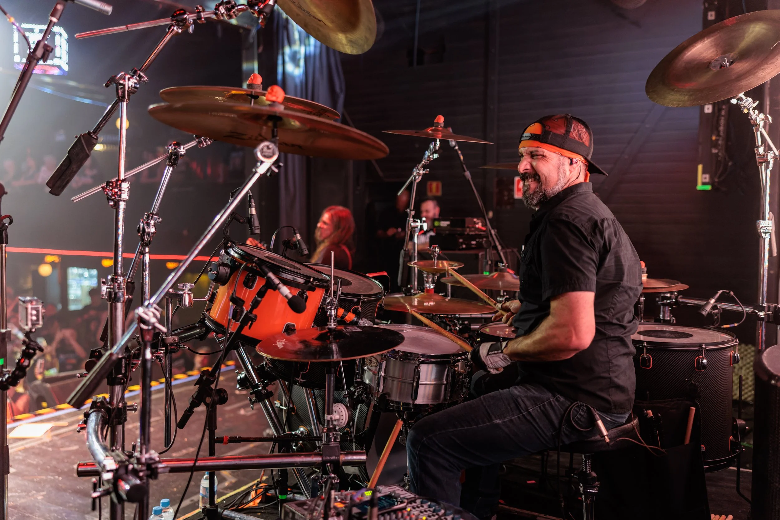 A male drummer with a beard and backwards baseball cap, playing a drum set on stage during a concert. The stage is filled with drums, cymbals, and other musical equipment, with stage lights illuminating the scene, and audience members visible in the 