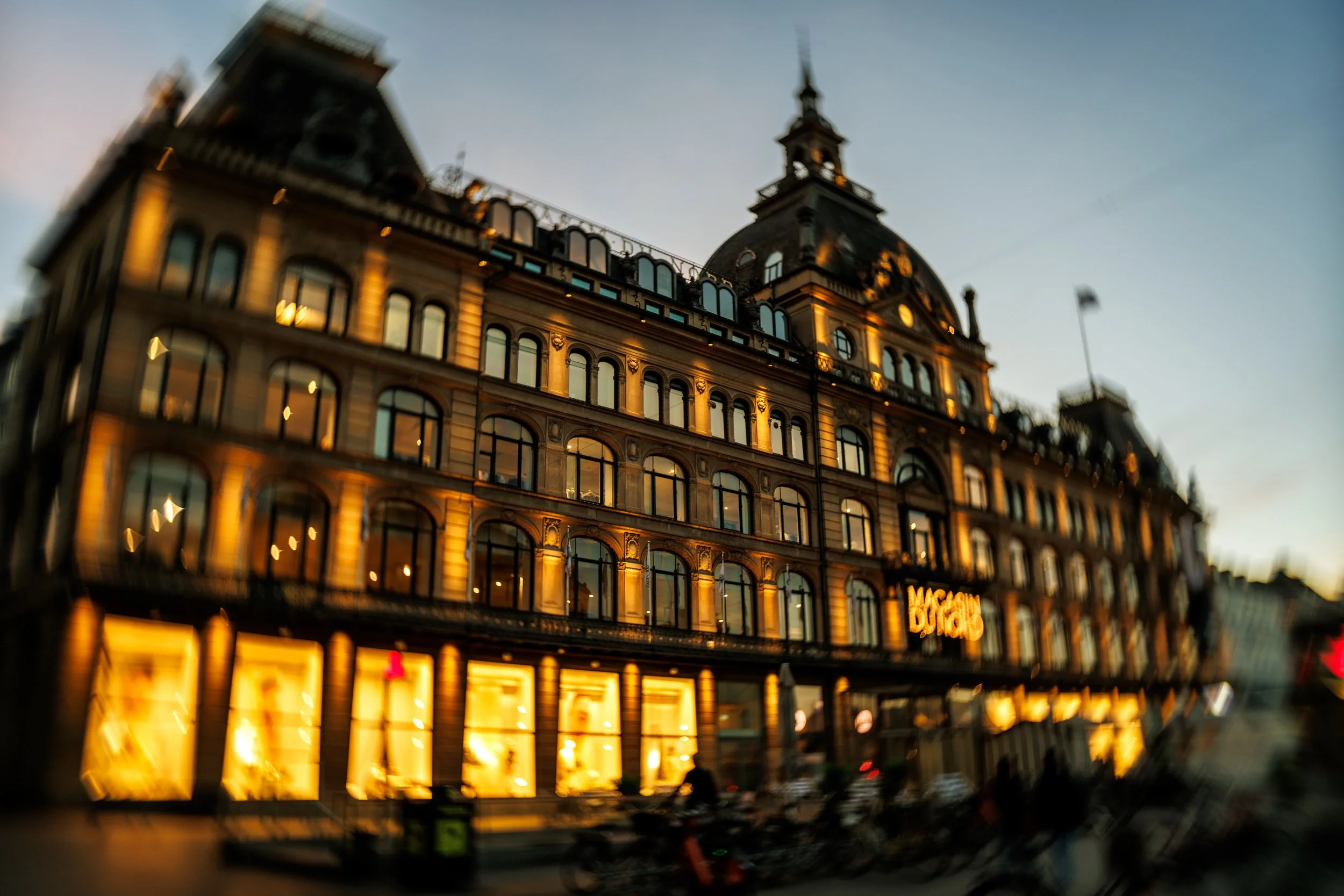 A multi-story building in the evening with lit windows, a prominent clock tower, and the Macy's sign illuminated on the lower level.