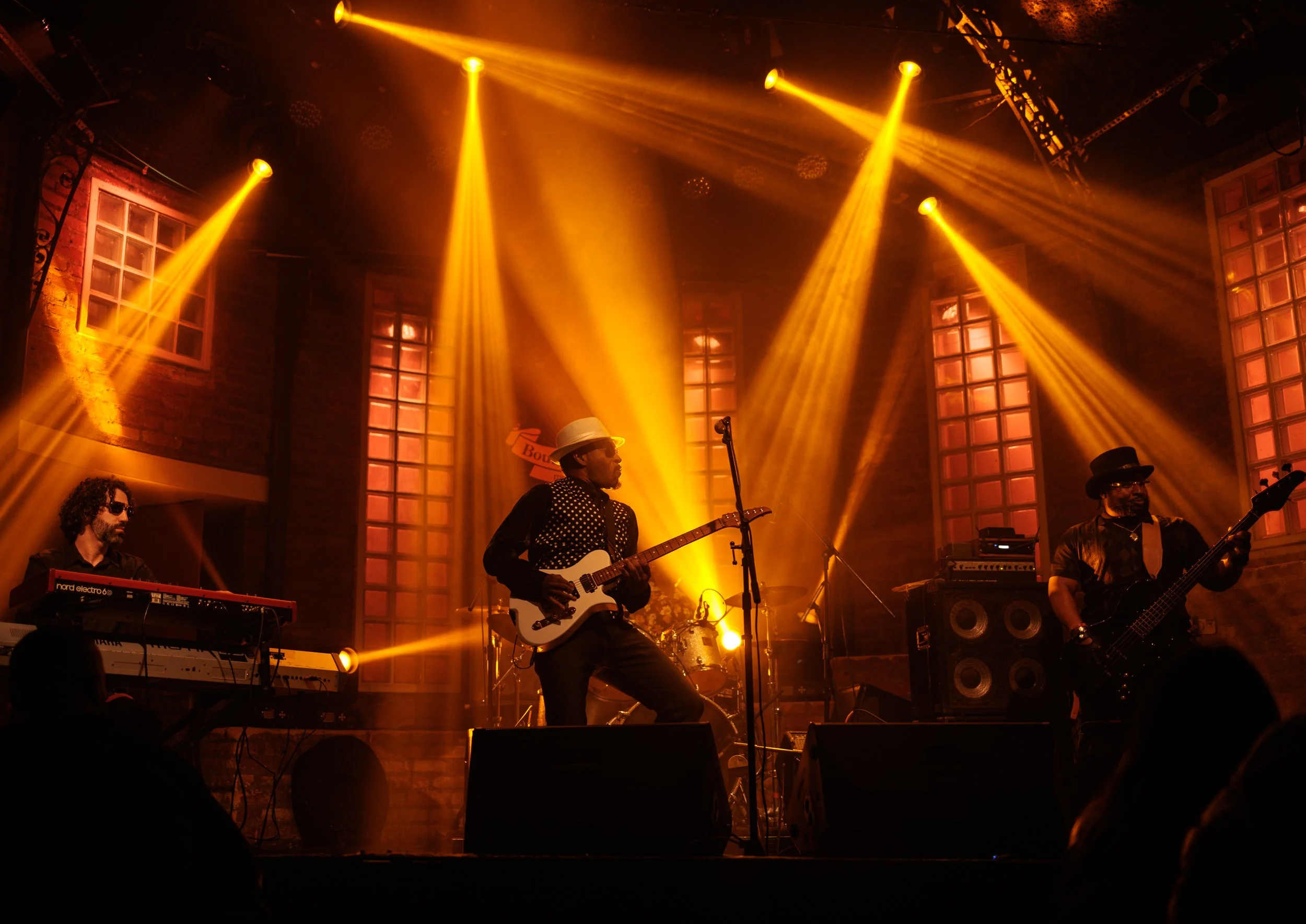Musicians performing on stage with yellow stage lights, brick wall background, and large windows. The band includes a keyboard player, a guitarist, and a bassist.
