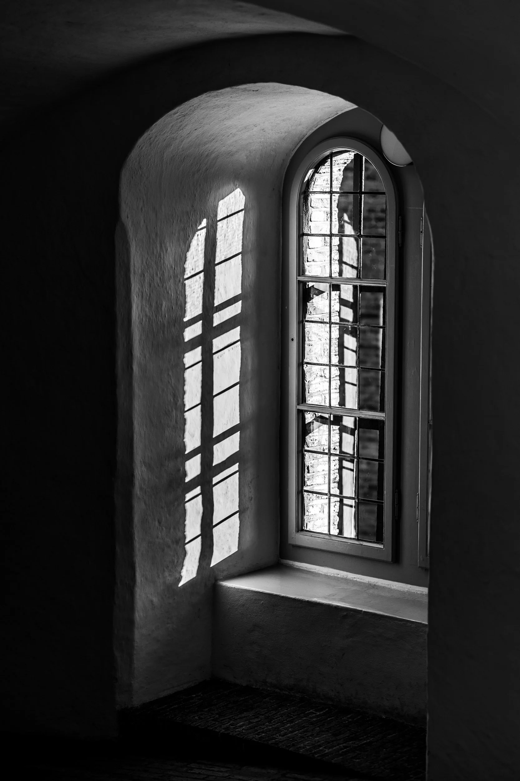 Black and white photo of a small arched window with sunlight casting a grid pattern of shadows on the wall inside a room with textured walls and wooden floors.