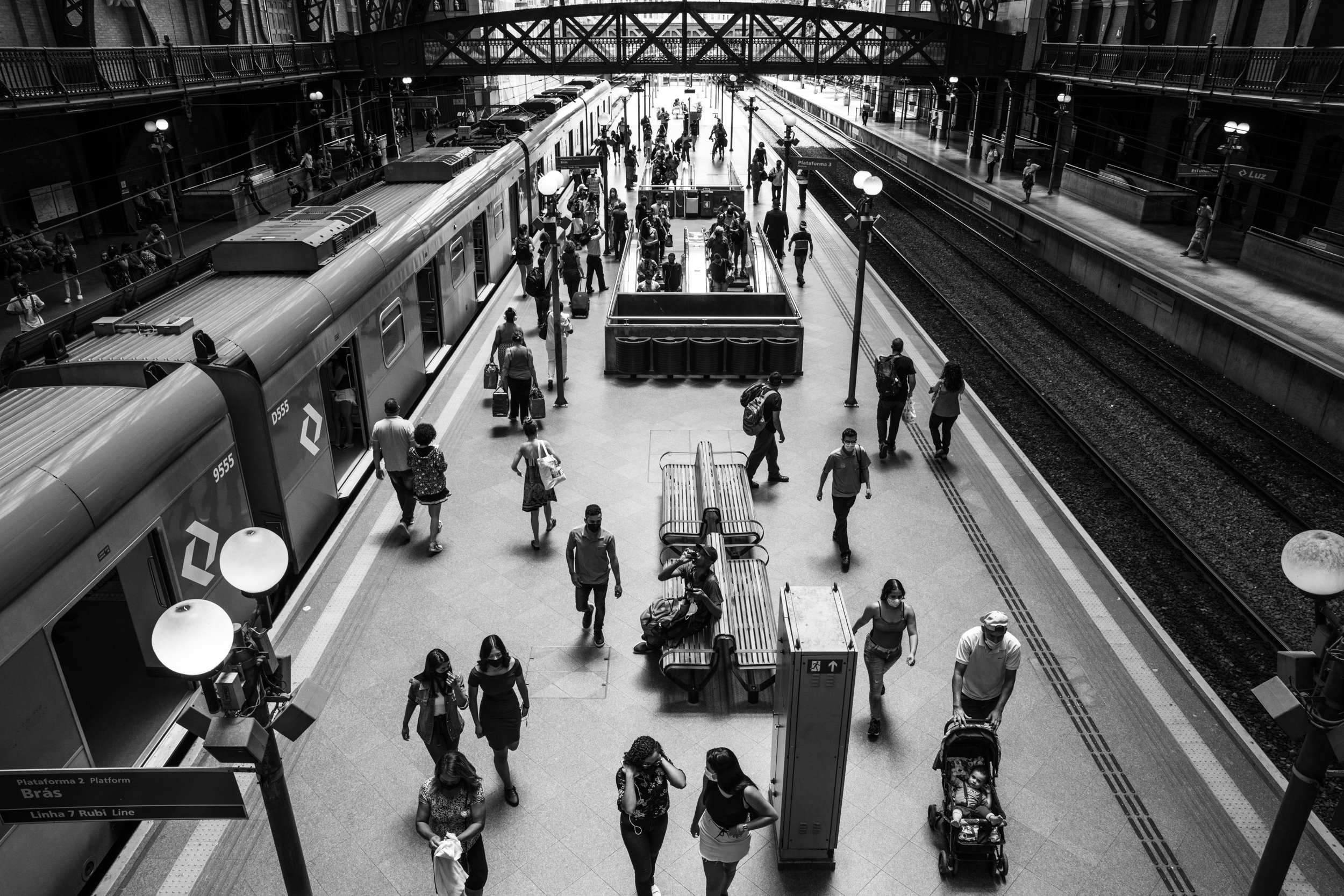 People walking inside a busy train station, some carrying luggage. A train is on the left side, and there are multiple platforms and tracks visible. The station has high ceilings and an overhead walkway.