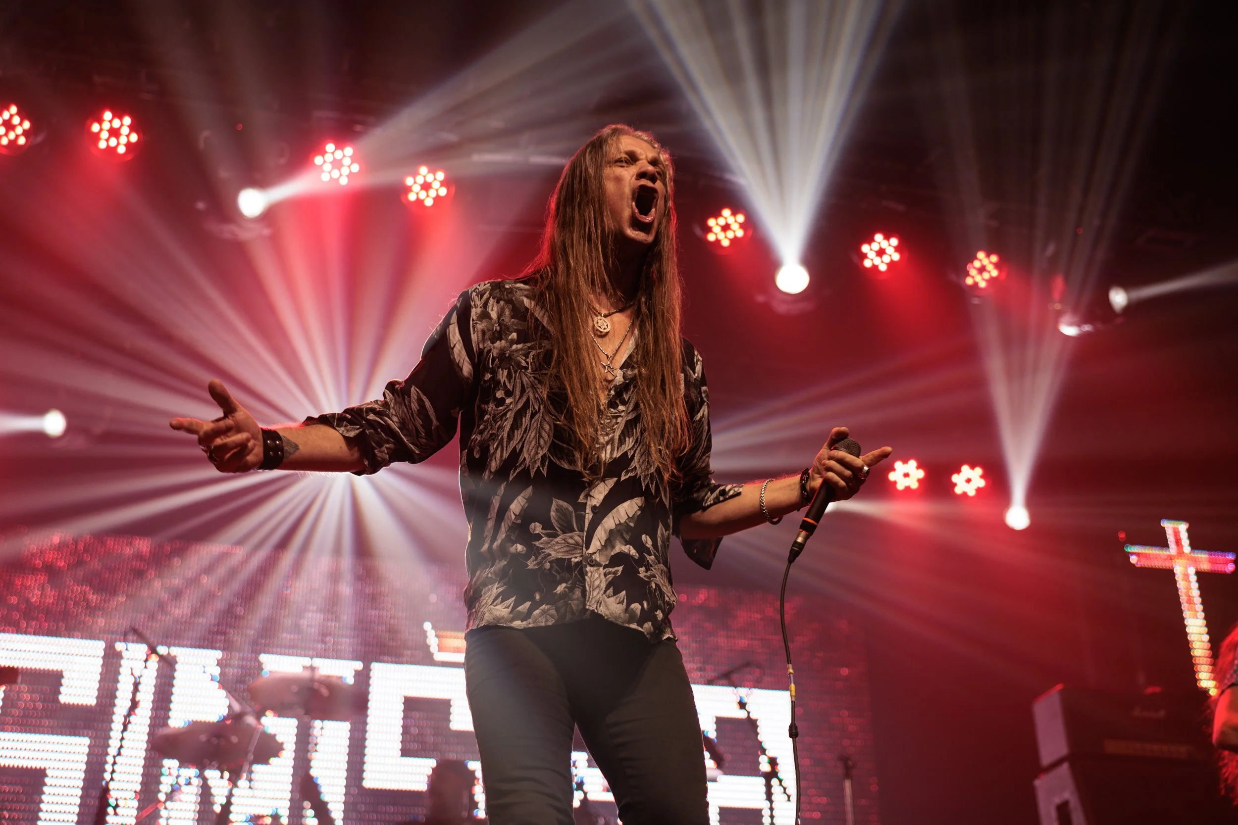 Male singer with long hair and patterned shirt performing passionately on stage with colorful stage lights and a large LED screen in the background.