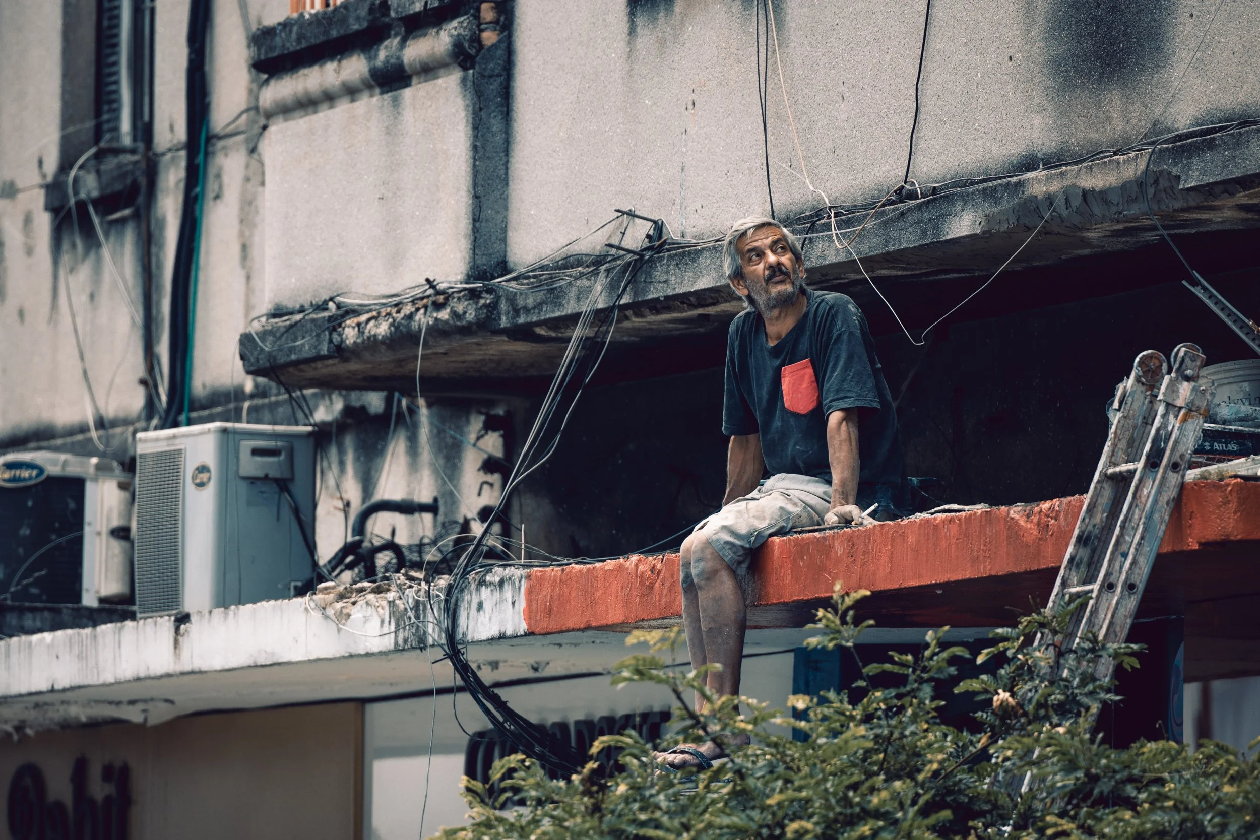 An older man with gray hair and a beard sitting on the edge of a building under renovation, surrounded by construction wires and tools, with a ladder nearby and air conditioning units on the building wall.