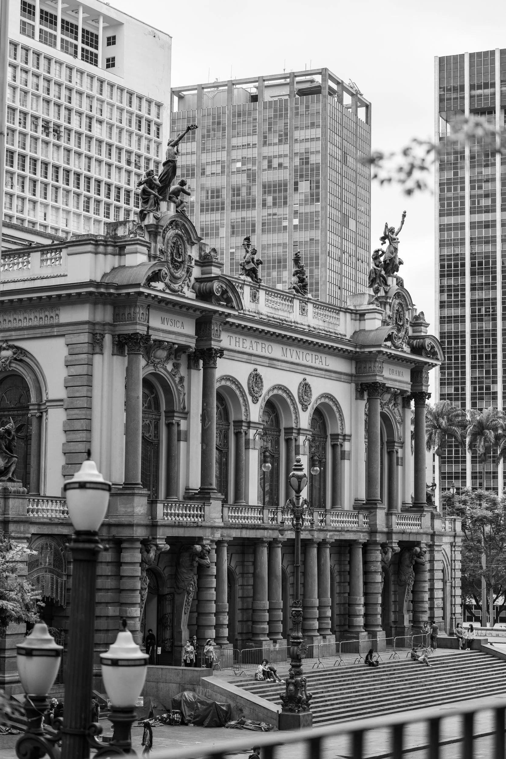 A black and white photo of an ornate historic building with statues on the roof, labeled 'Theatro Municipal' in Portuguese, with modern high-rise buildings in the background. People are gathered on the steps and around the entrance.