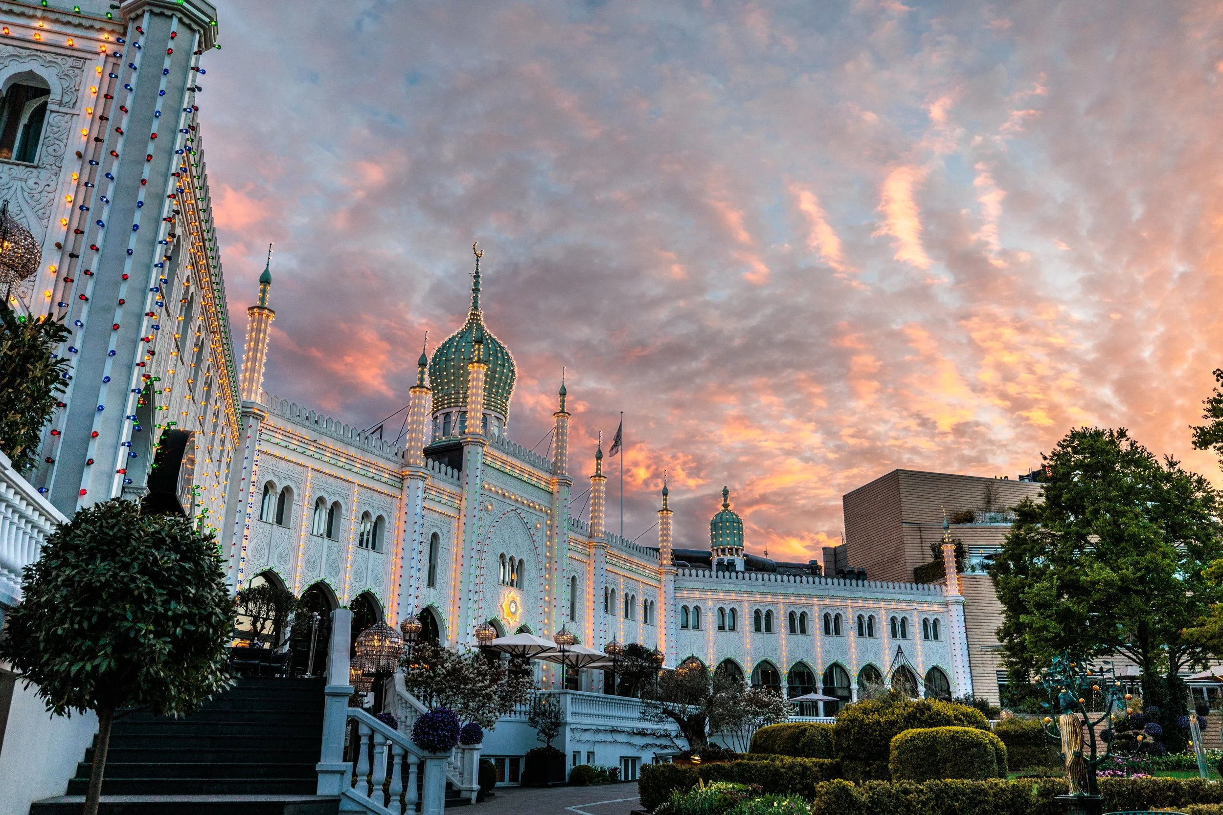 A white castle-like building decorated with colorful string lights under a pink and blue cloudy sky at sunset, with trees and plants in the foreground.