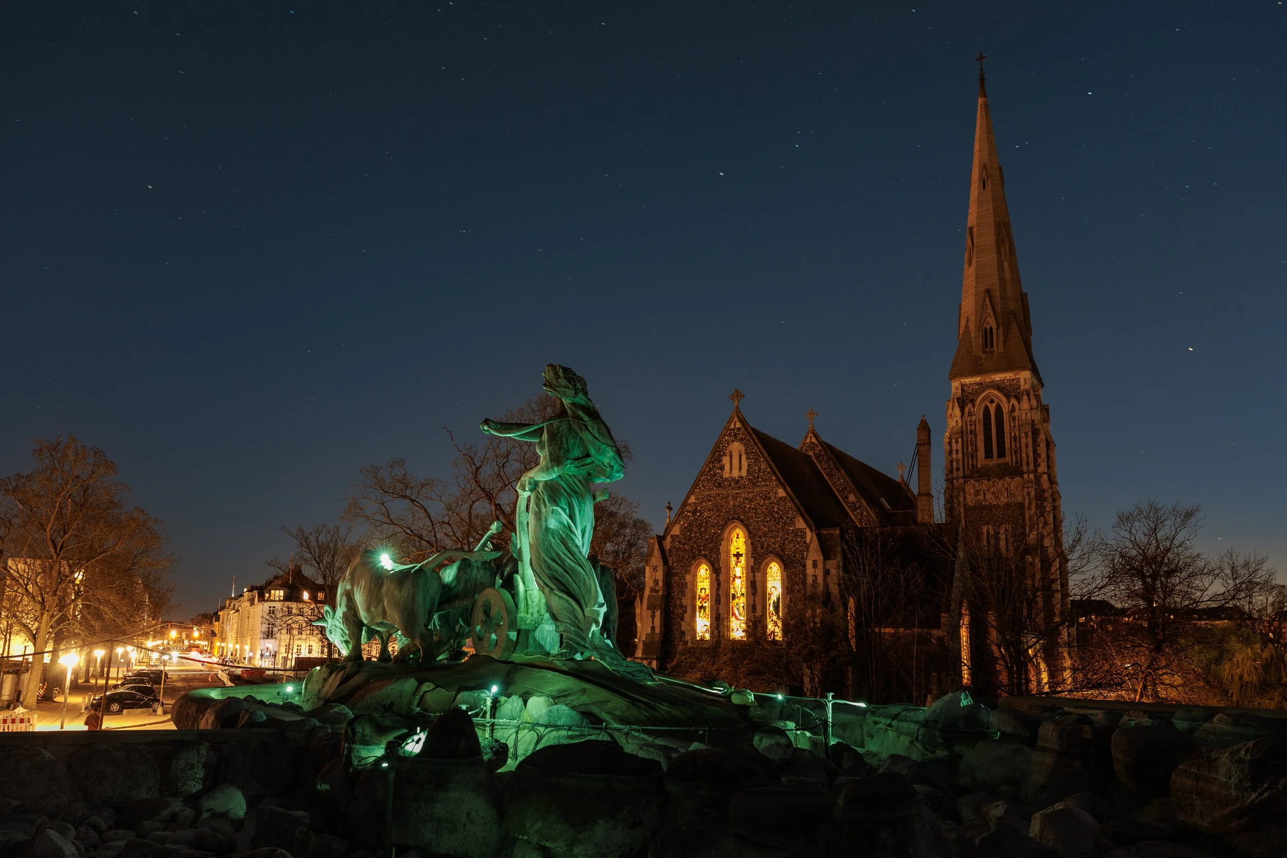 Night view of a church with a tall steeple and stained glass windows, illuminated behind a bronze statue of a chariot with horses and a figure, with starry sky above and trees and streetlights in foreground.