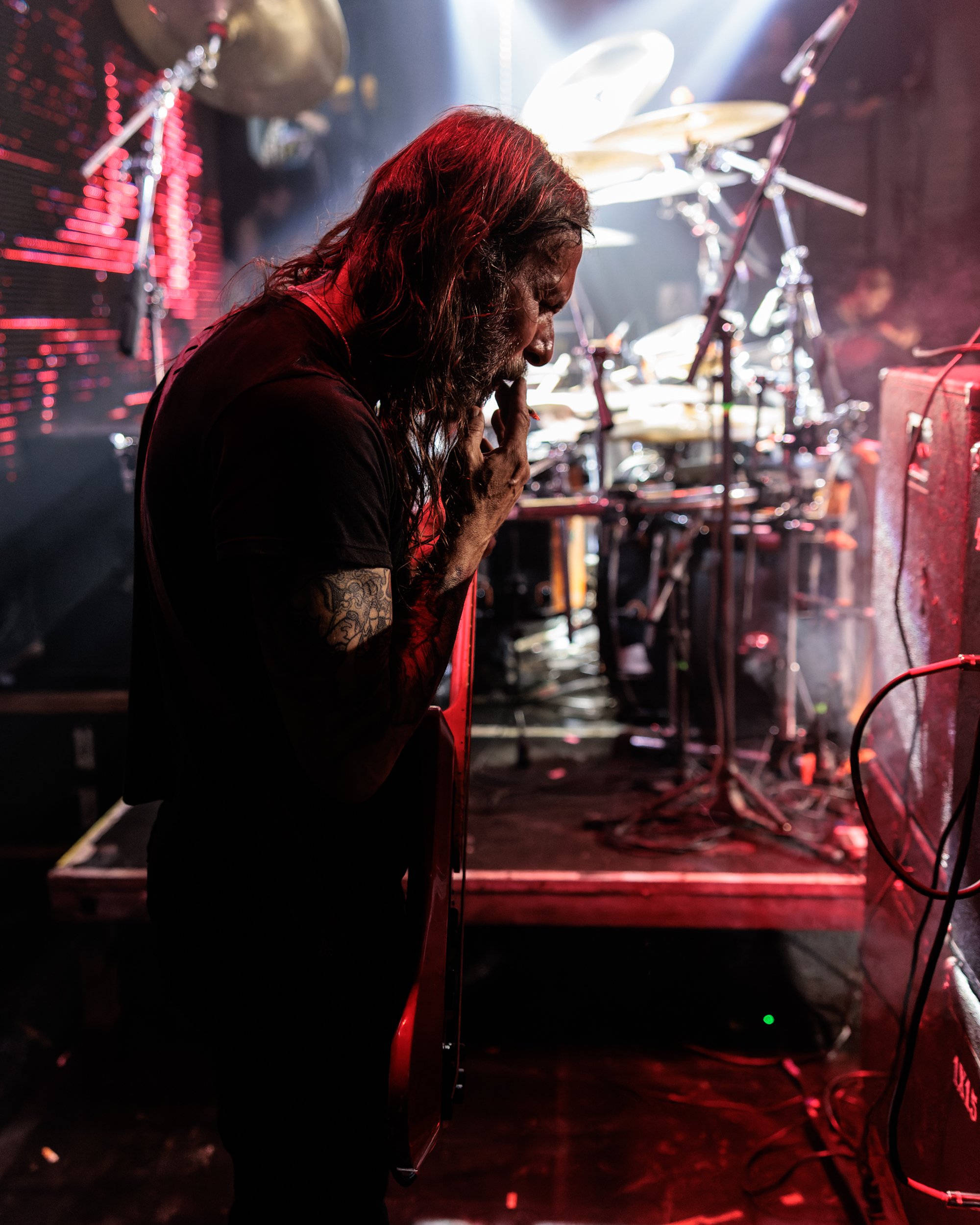 A man with long hair and tattoos on his arm, standing in front of a drum set on a dark stage. The man appears to be praying or contemplating, illuminated by stage lights with a red hue.