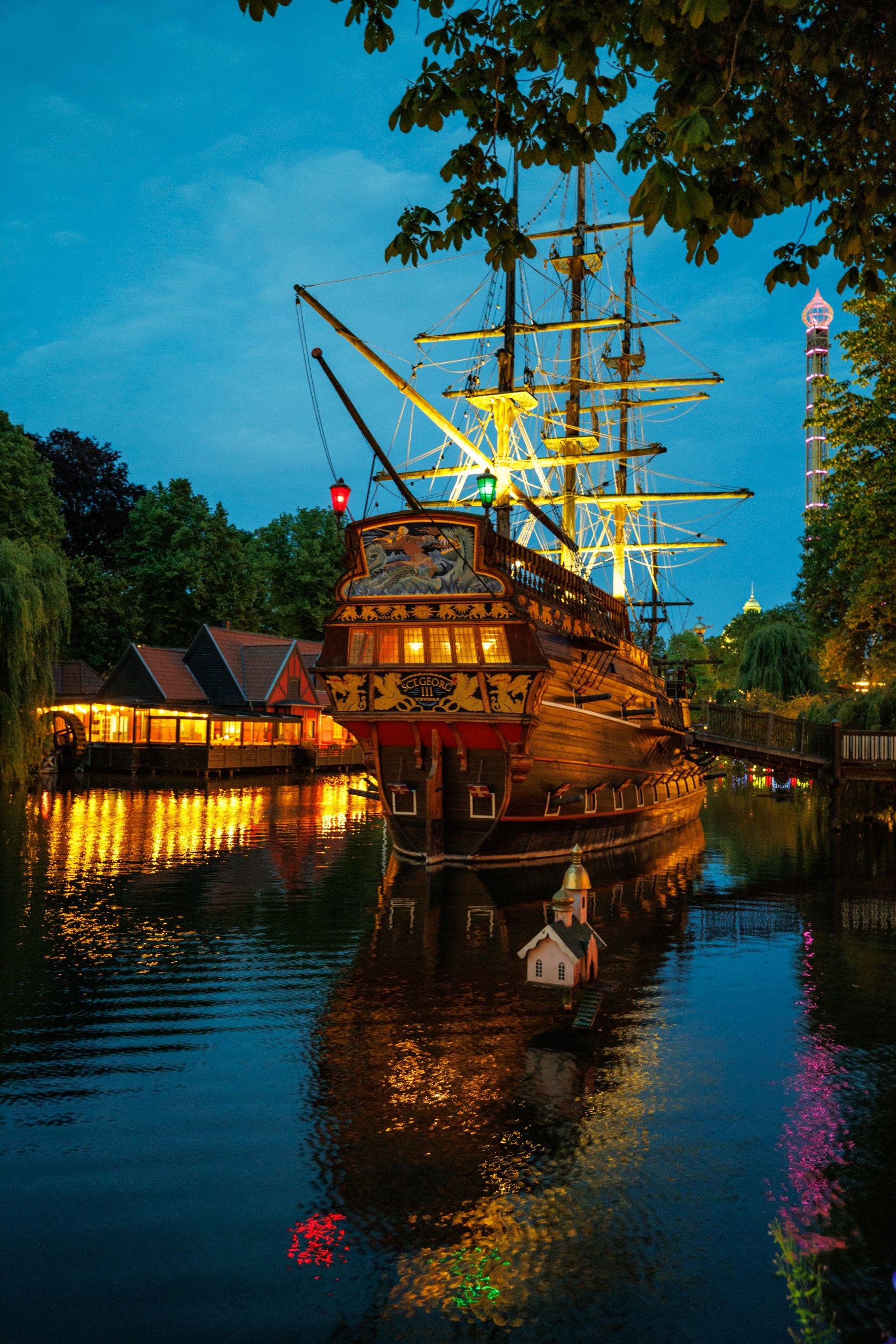 A historic wooden sailing ship with illuminated masts and rigging, floating on a calm river with reflections of colorful lights, surrounded by trees and a small boat house, during evening or night.