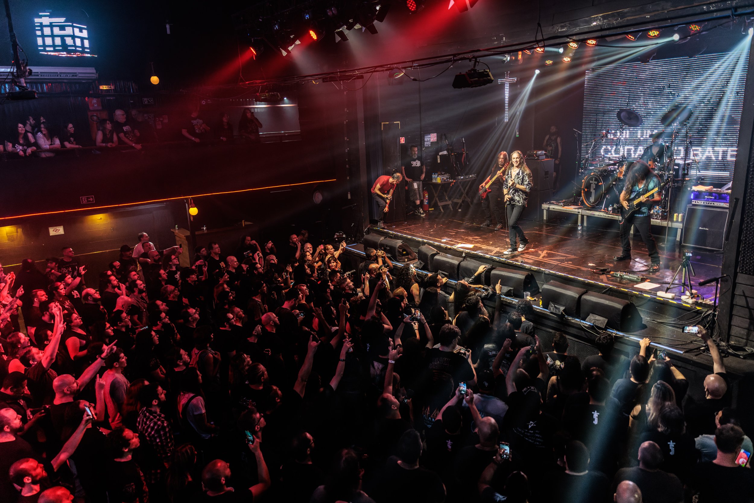 A live music concert with a band performing on stage, an audience cheering and taking photos, and colorful stage lighting in a dark indoor venue.