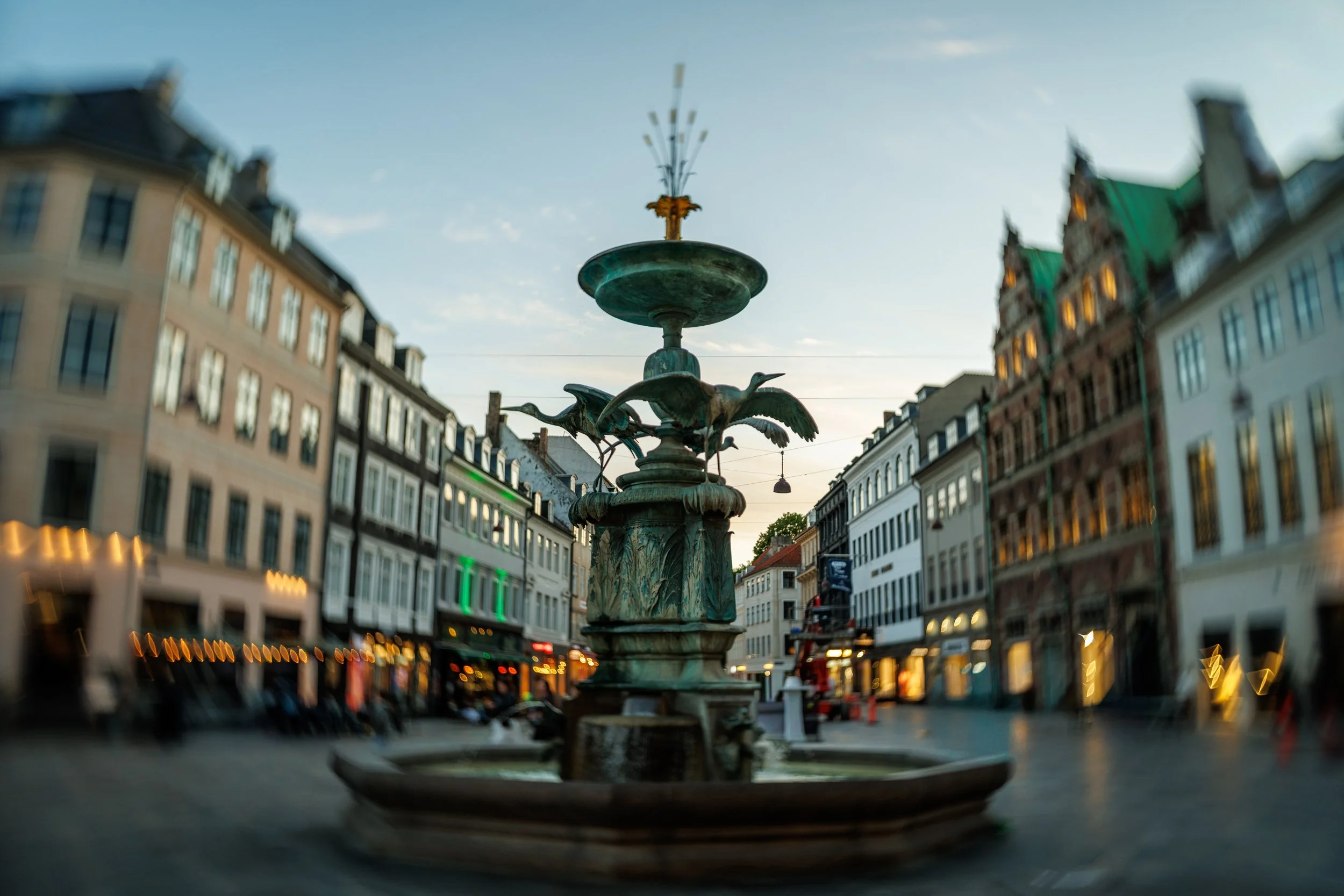 An ornate fountain with sculptures of birds in the middle of a pedestrian street in a European city, surrounded by multi-story buildings with varying architecture, illuminated by streetlights at dusk.