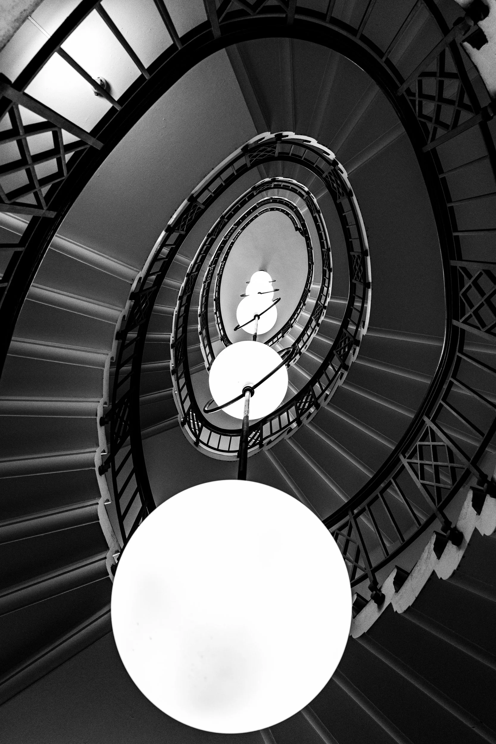 A black and white photo taken from the bottom of a spiral staircase looking up, with illuminated globe lights hanging along the center.