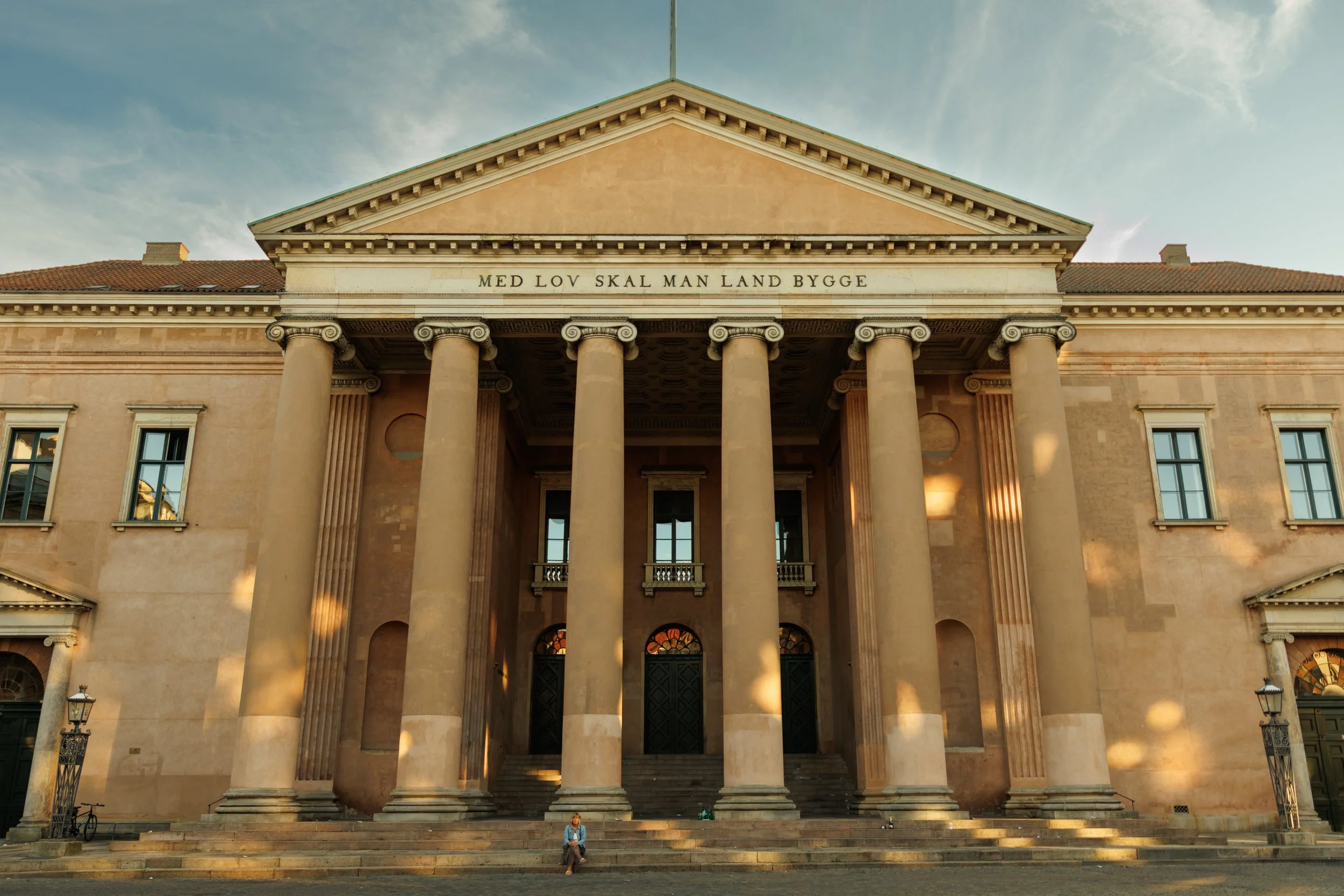 A neoclassical building with large columns, a triangular pediment with an inscription, and a person sitting on the stairs in front.