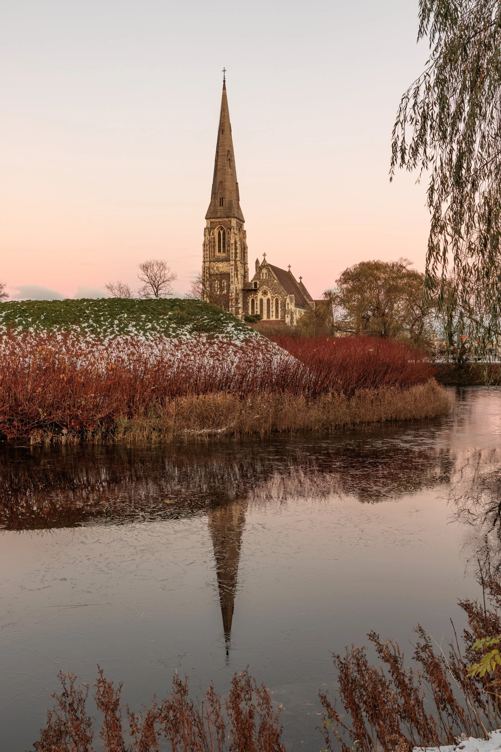 A church with a tall steeple situated near a body of water, with its reflection visible in the water, surrounded by trees and bushes, under a pastel-colored sky during sunset or sunrise.