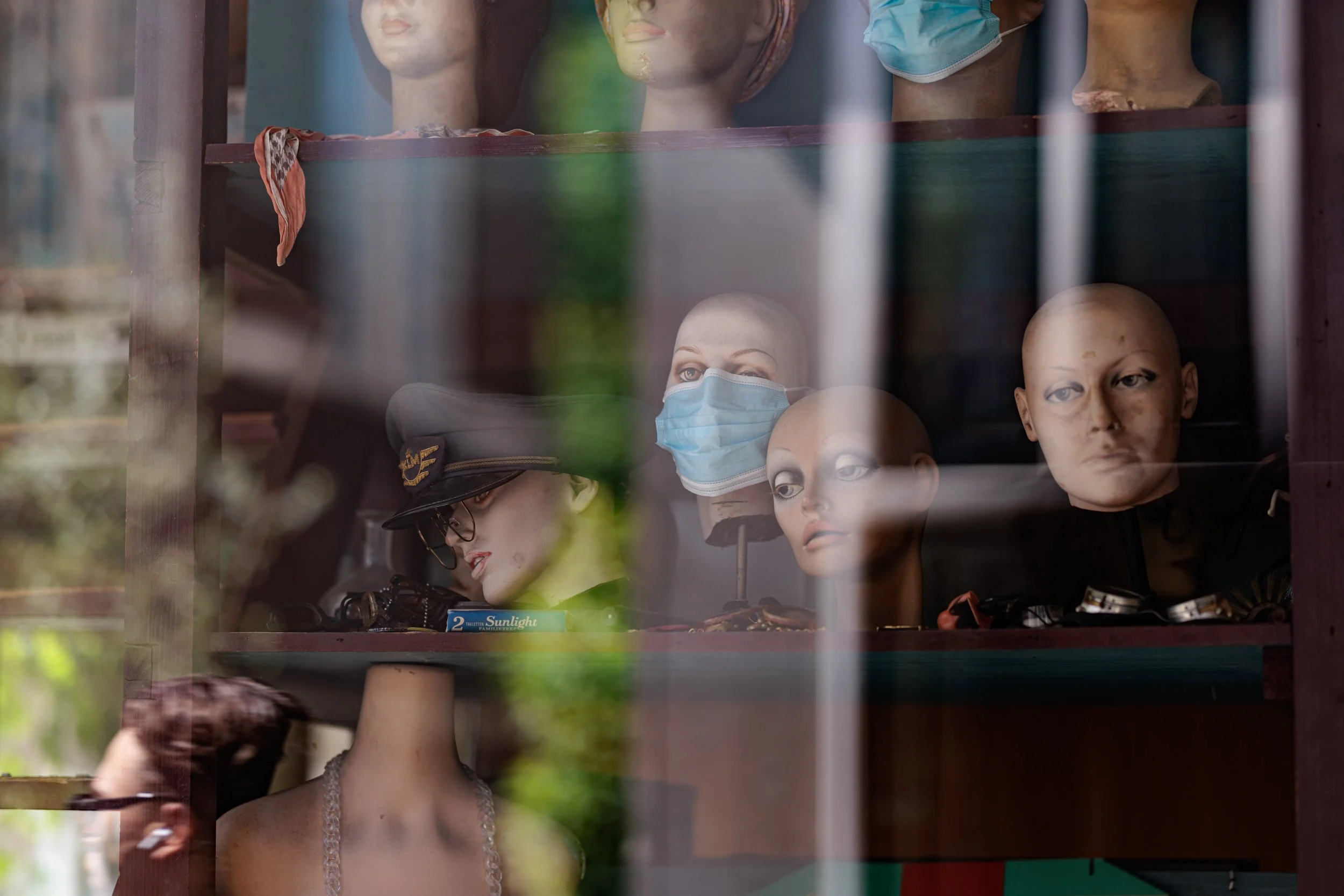 Display window with mannequin heads, some wearing face masks, and assorted accessories behind glass.