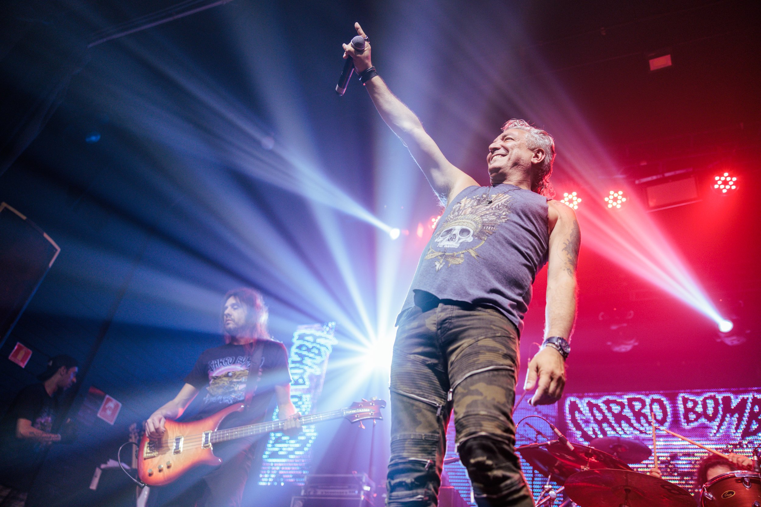 A male singer with gray hair, wearing a sleeveless shirt with a skull graphic, holding a microphone and pointing upward, performing on stage with band members and colorful stage lights in the background.