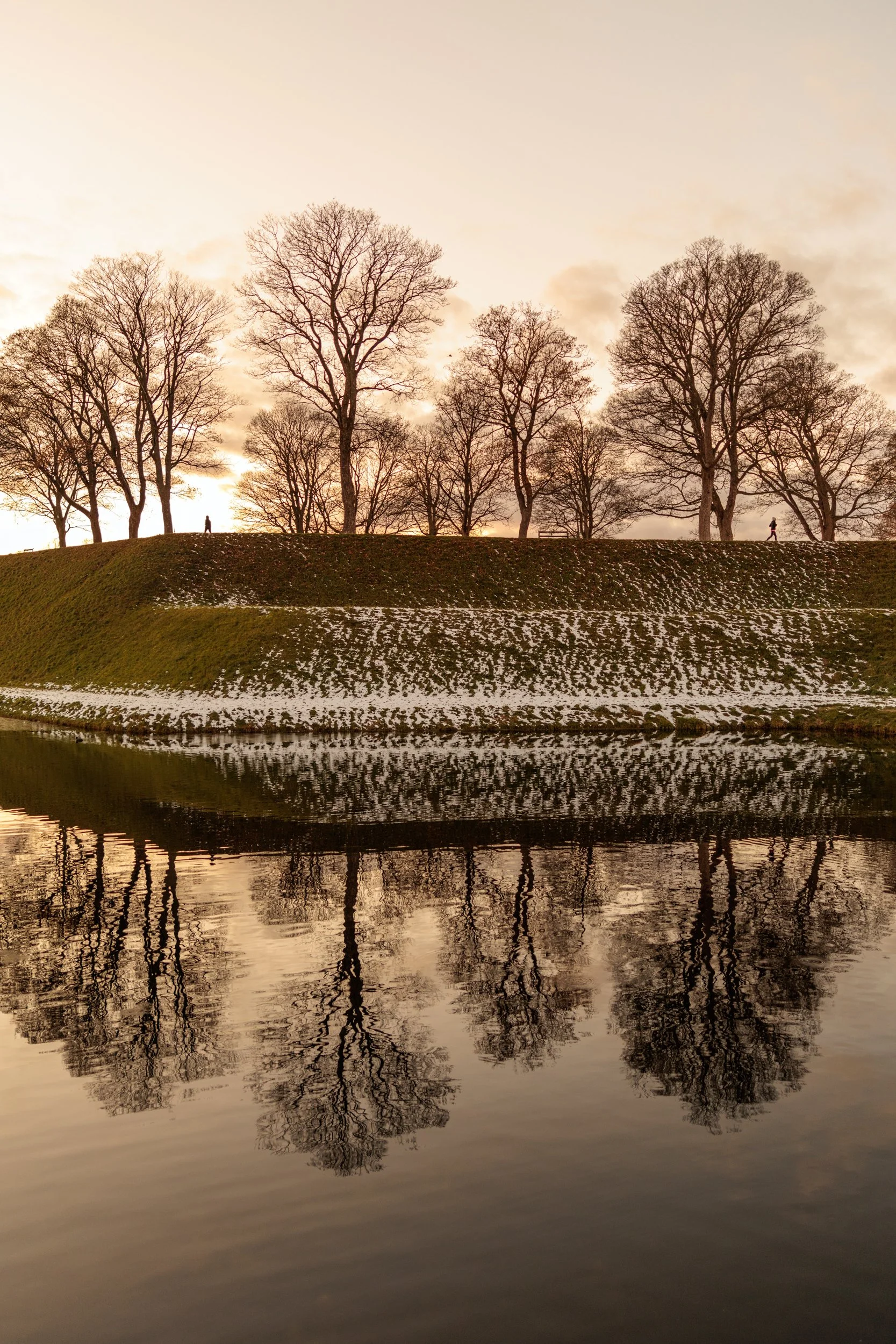 Sunset over a hill with leafless trees and their reflection in a calm body of water, with snow patches on the grass.