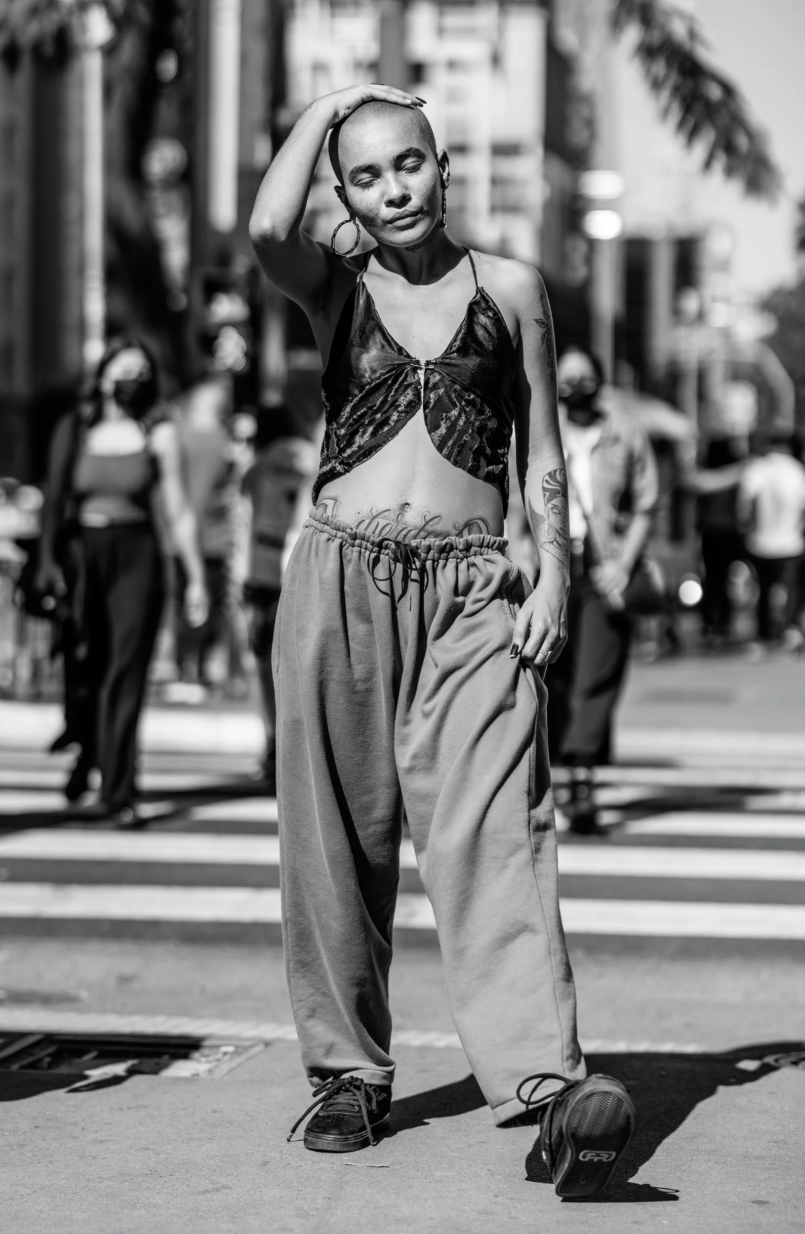 A black and white photo of a woman with a shaved head, wearing large hoop earrings, a top with a butterfly design, and loose pants. She is standing in the street with her eyes closed and hand resting on her head, with a background of people and build