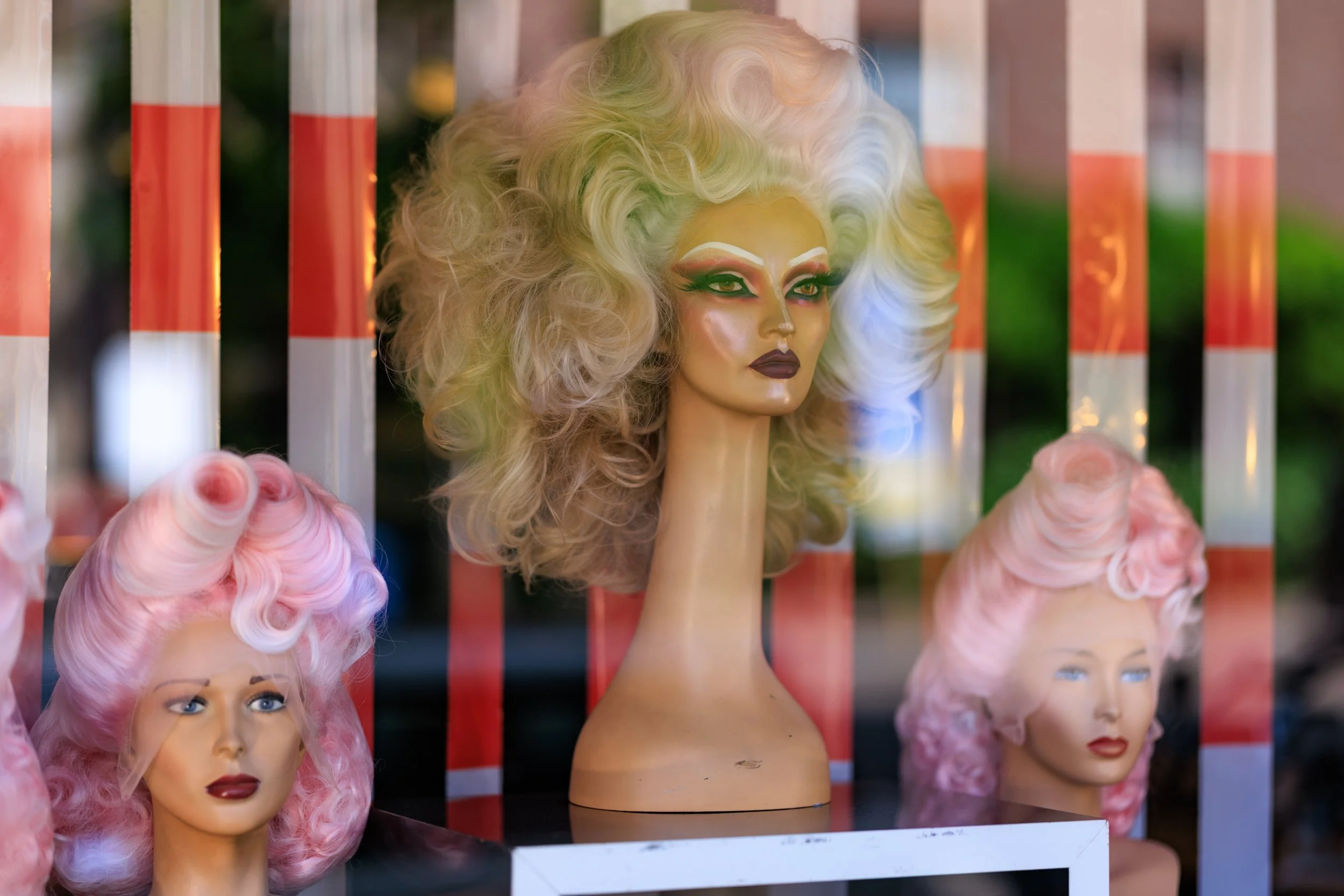 Display of various mannequin heads with colorful artistic makeup, wigs, and headpieces behind store glass.