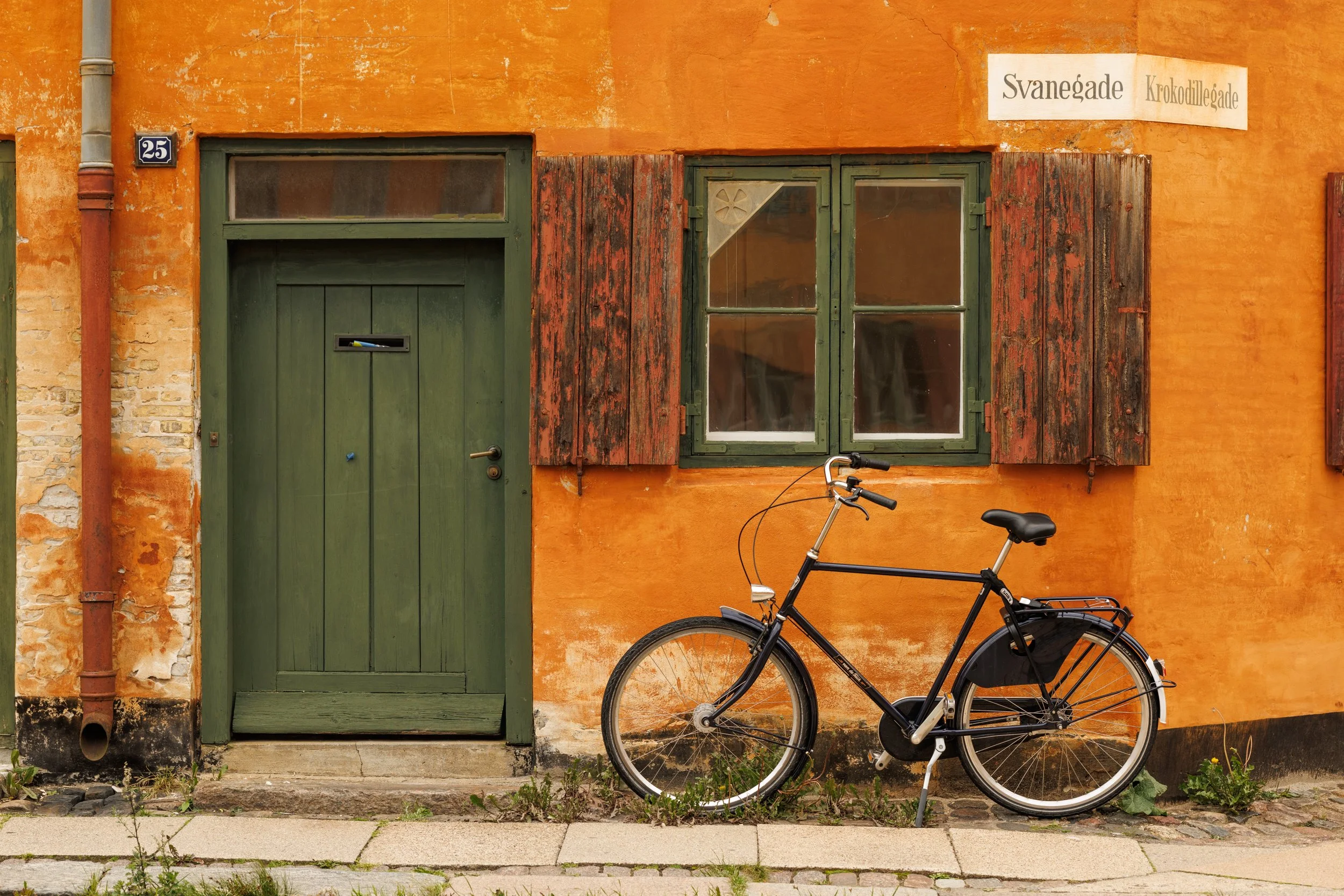 An orange building with a green door and window shutters, and a bicycle parked in front.